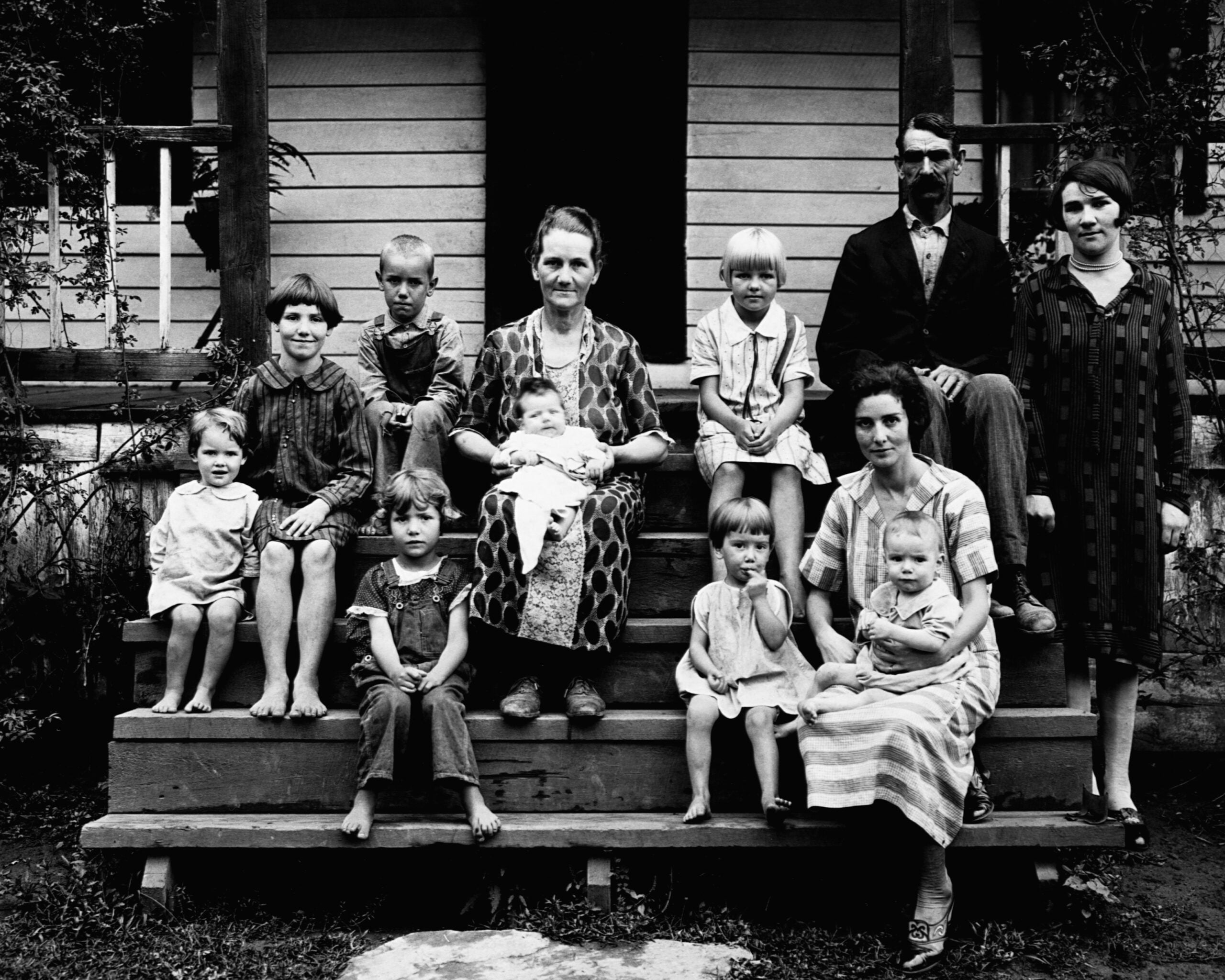 Working class families in the 1920s and 30s. Black and white photo of three adults and nine children sitting and standing on wooden steps in front of a house. The children are barefoot; two babies are held by women in the center and on the right.