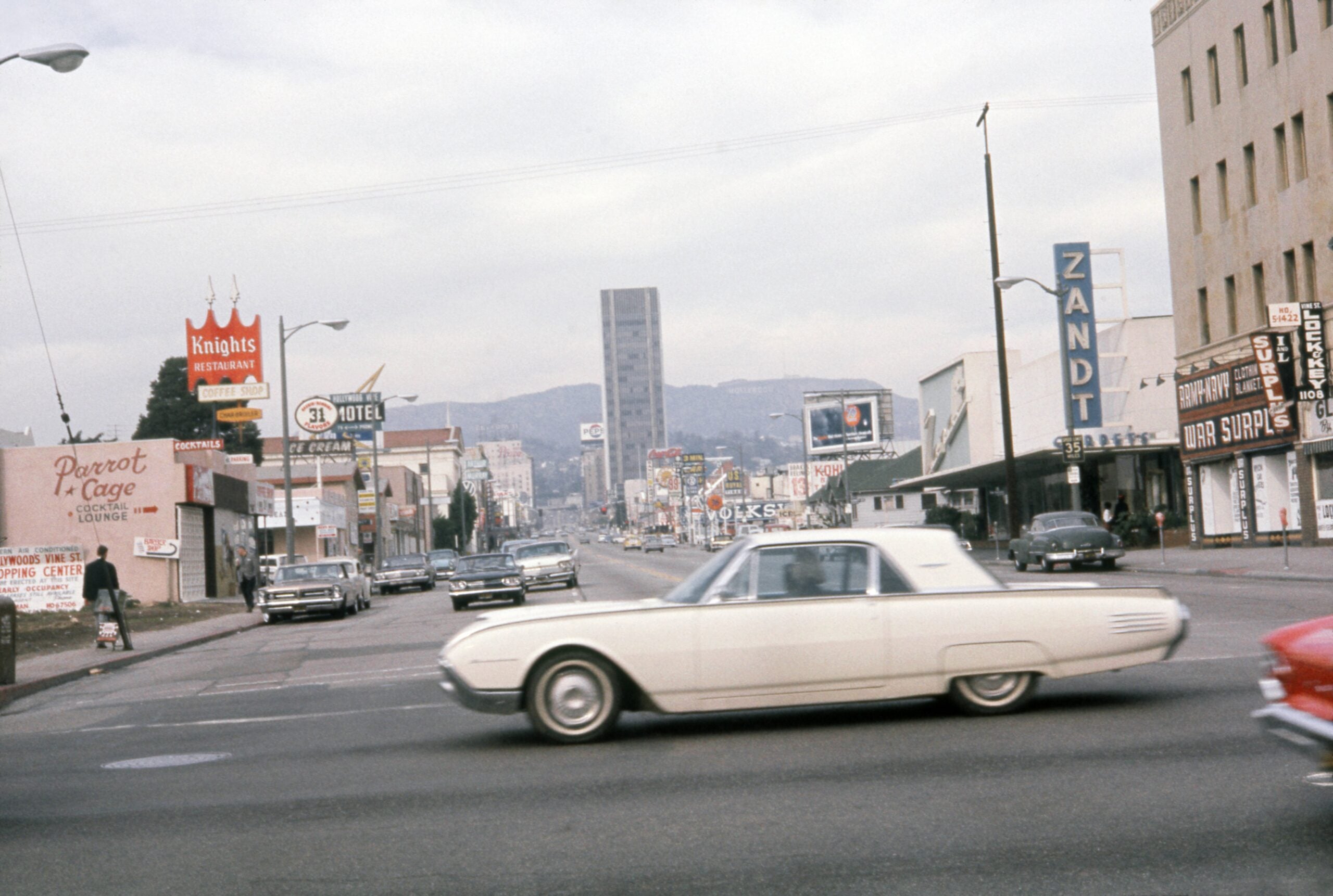 A vintage white car drives through a busy city intersection lined with classic cars, neon signs, and mid-century buildings under a cloudy sky.