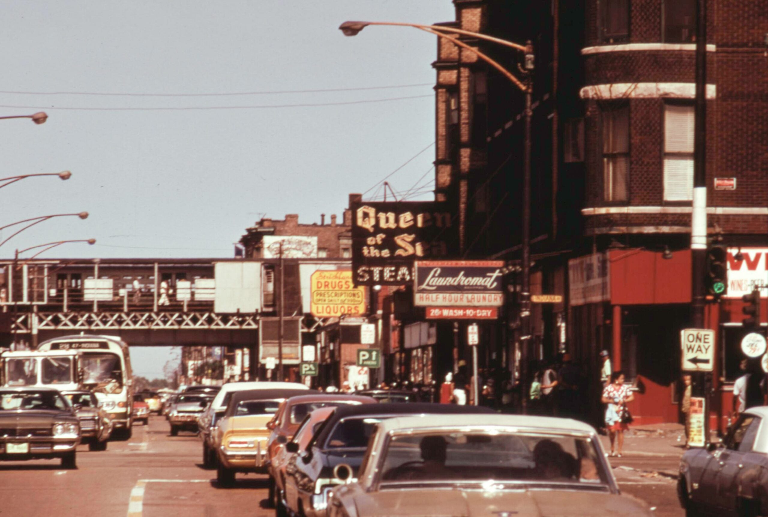 Busy city street scene with cars, buses, pedestrians, storefronts, vintage signs, and an elevated train track in the background. Brick buildings line the road under a clear daytime sky.