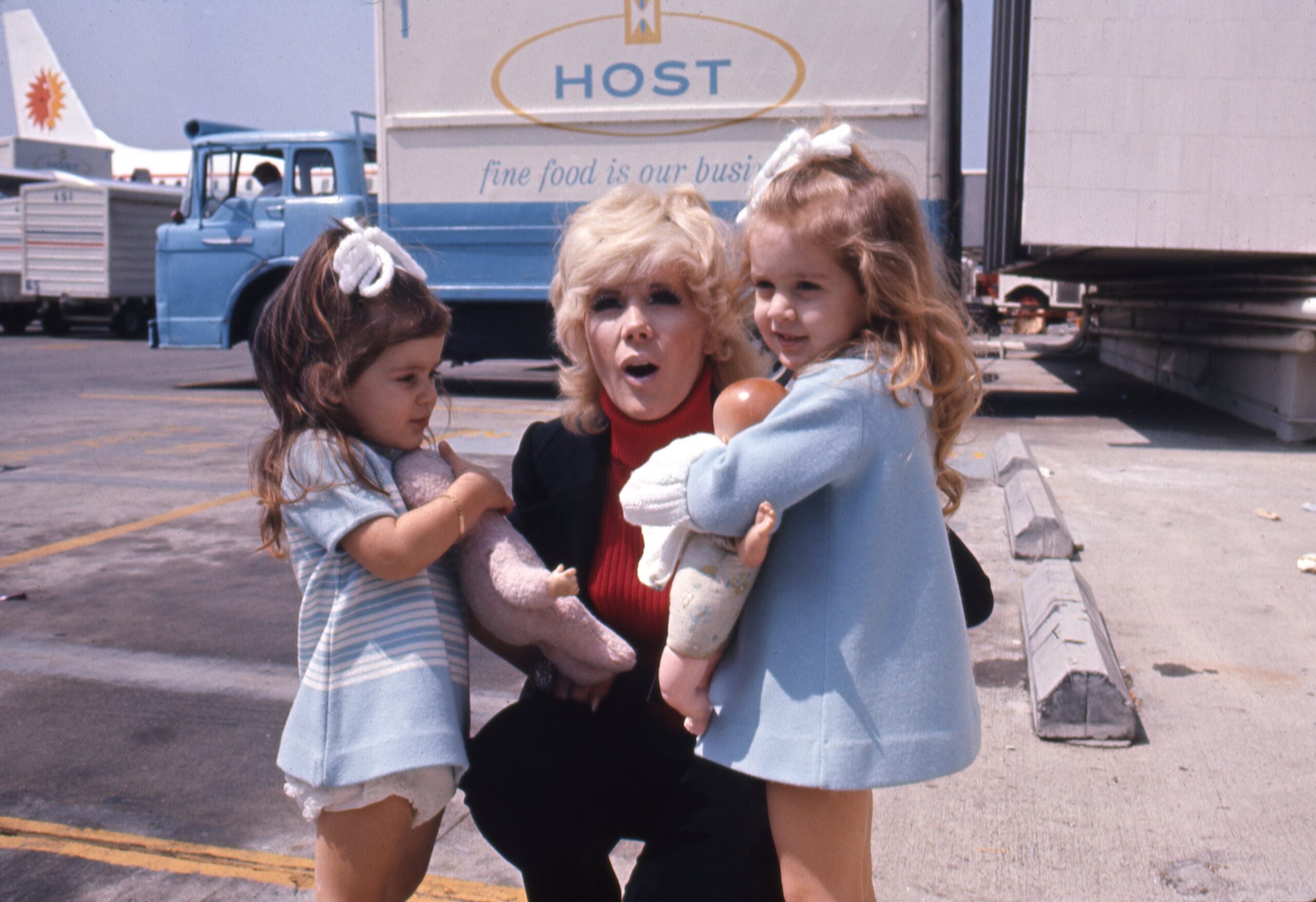 A woman kneels between two young girls holding dolls, all wearing blue outfits, on an airport tarmac with trucks and a HOST catering truck in the background.