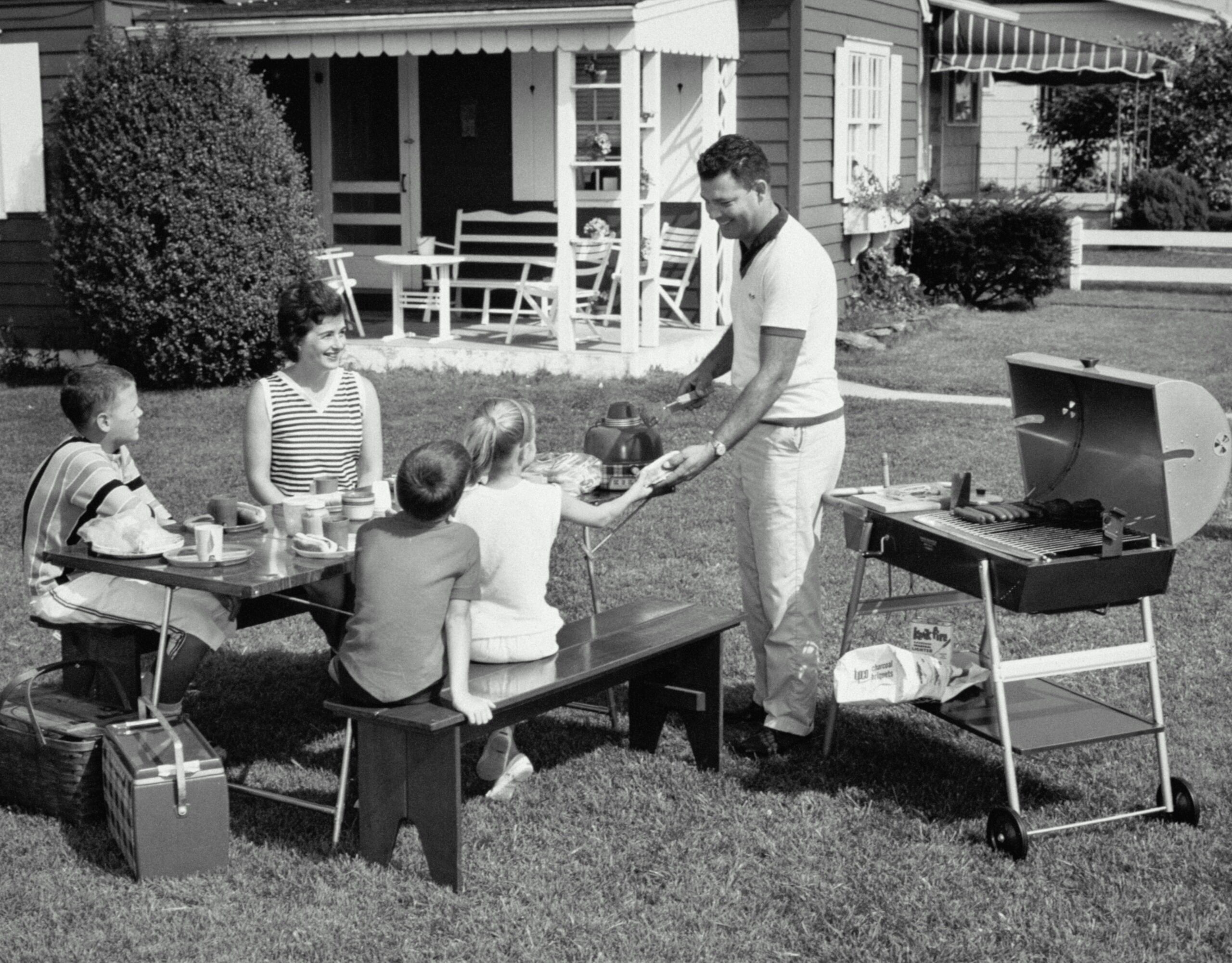 A man serves food to a woman and three children at a picnic table on a lawn, with a grill and house in the background. The scene suggests a family barbecue in a suburban yard.