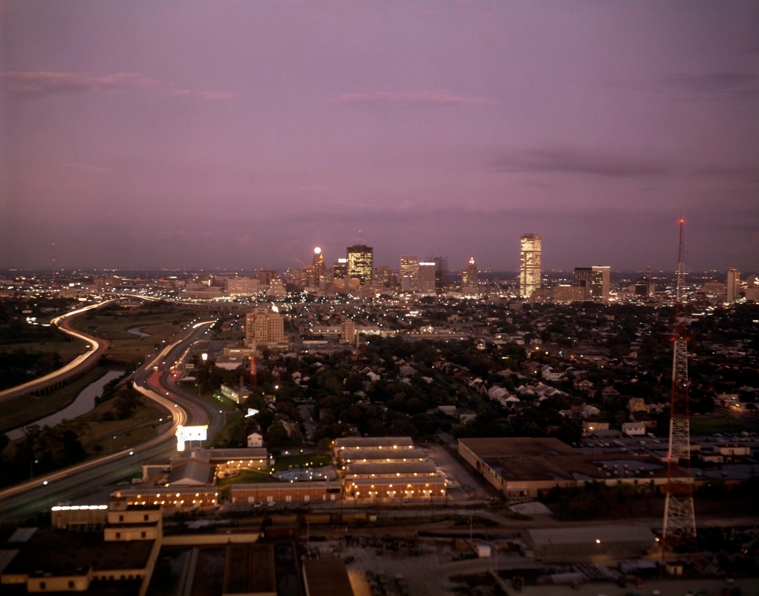City skyline at dusk with lit buildings, highways, and a river. The sky is a soft purple, and lights are visible throughout the city and streets. Industrial buildings and a radio tower are in the foreground.