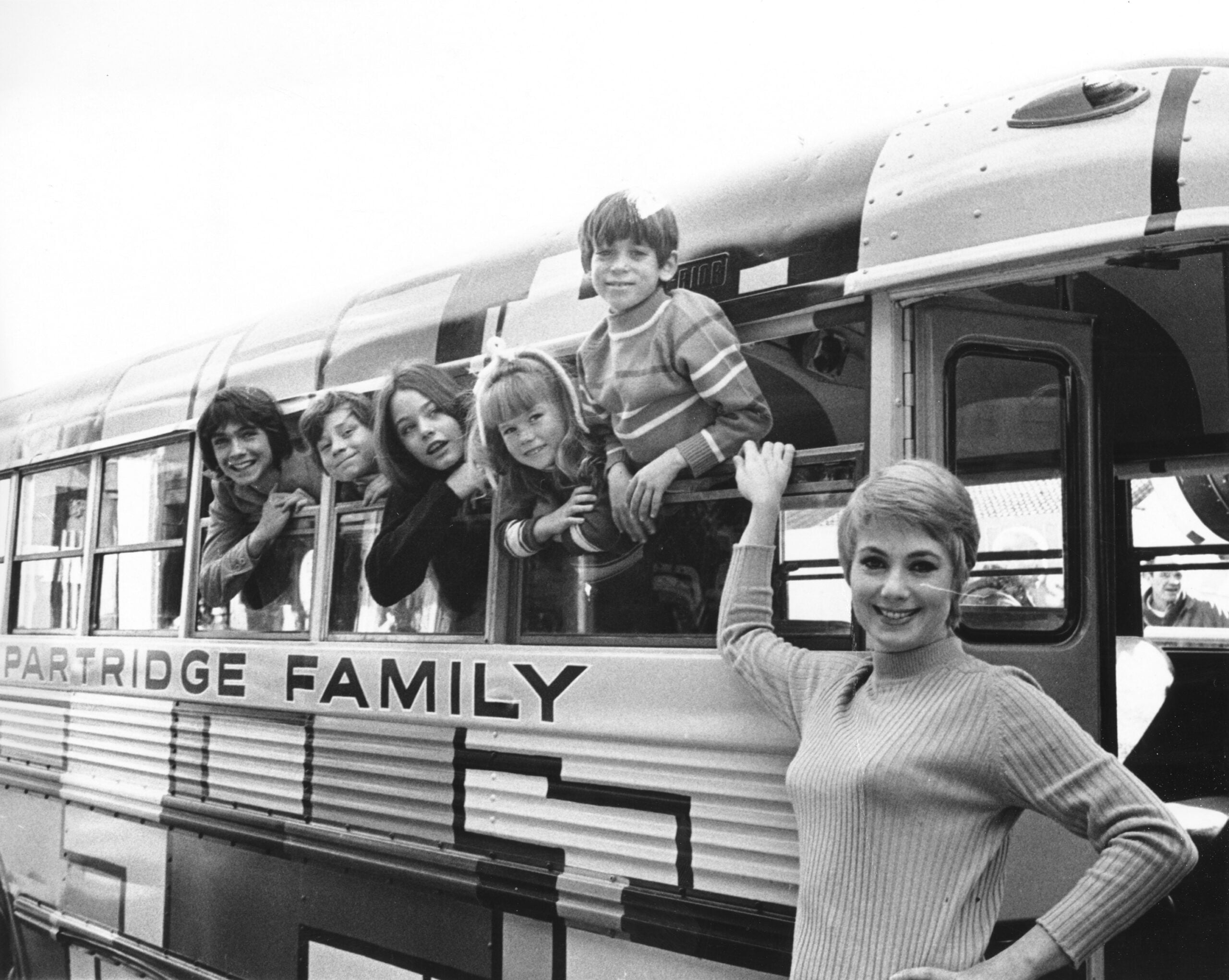 Five smiling people lean out bus windows labeled “Partridge Family,” while a woman stands outside, hand on the bus. The group appears cheerful and close, posing for the camera in this black-and-white photo.