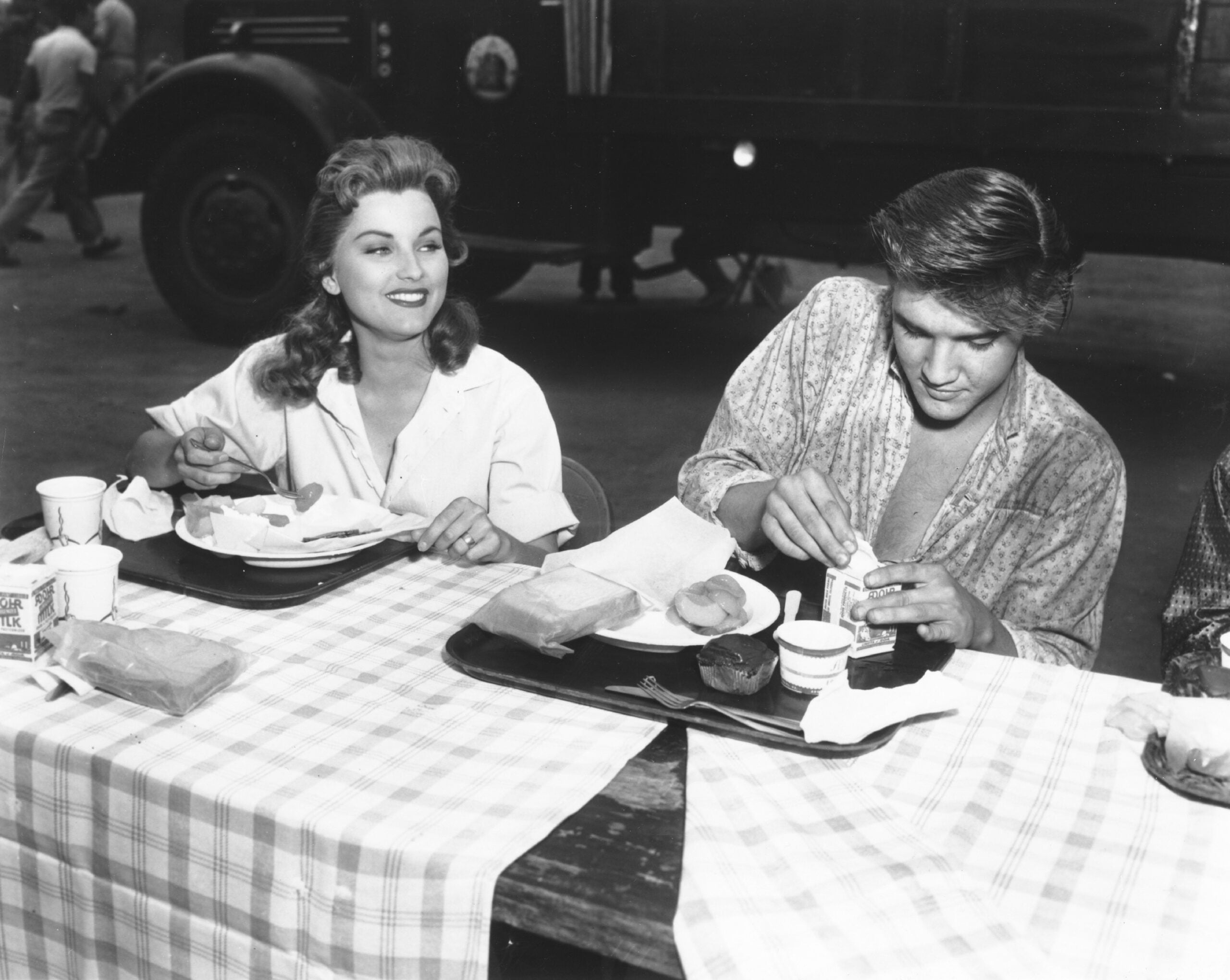A smiling woman and a man eat lunch together at a checkered table outdoors, using trays and paper cups. A truck and people are visible in the background.