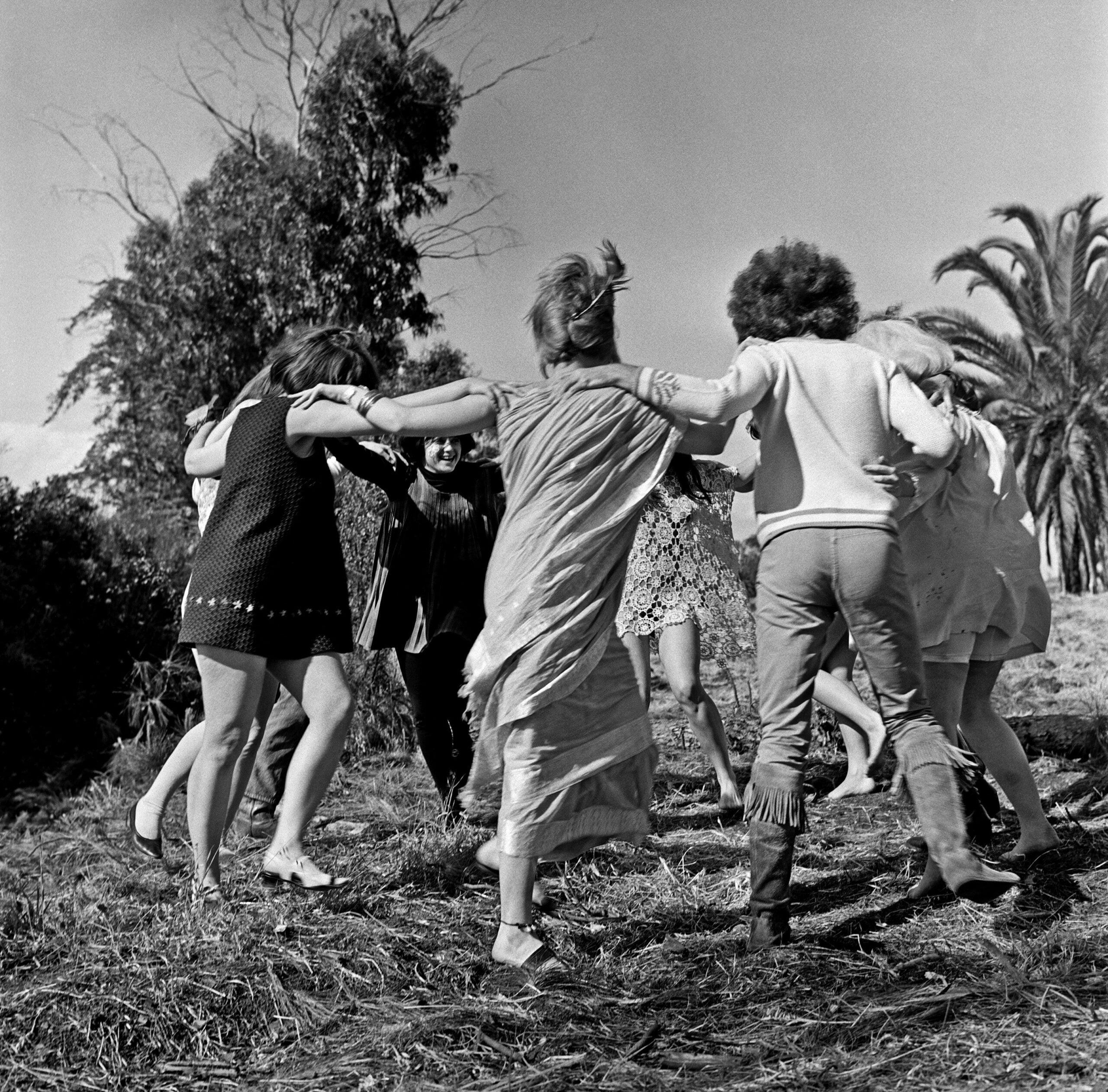 A group of people stand outdoors, holding hands in a circle and dancing together on grass, with trees and palm trees in the background.