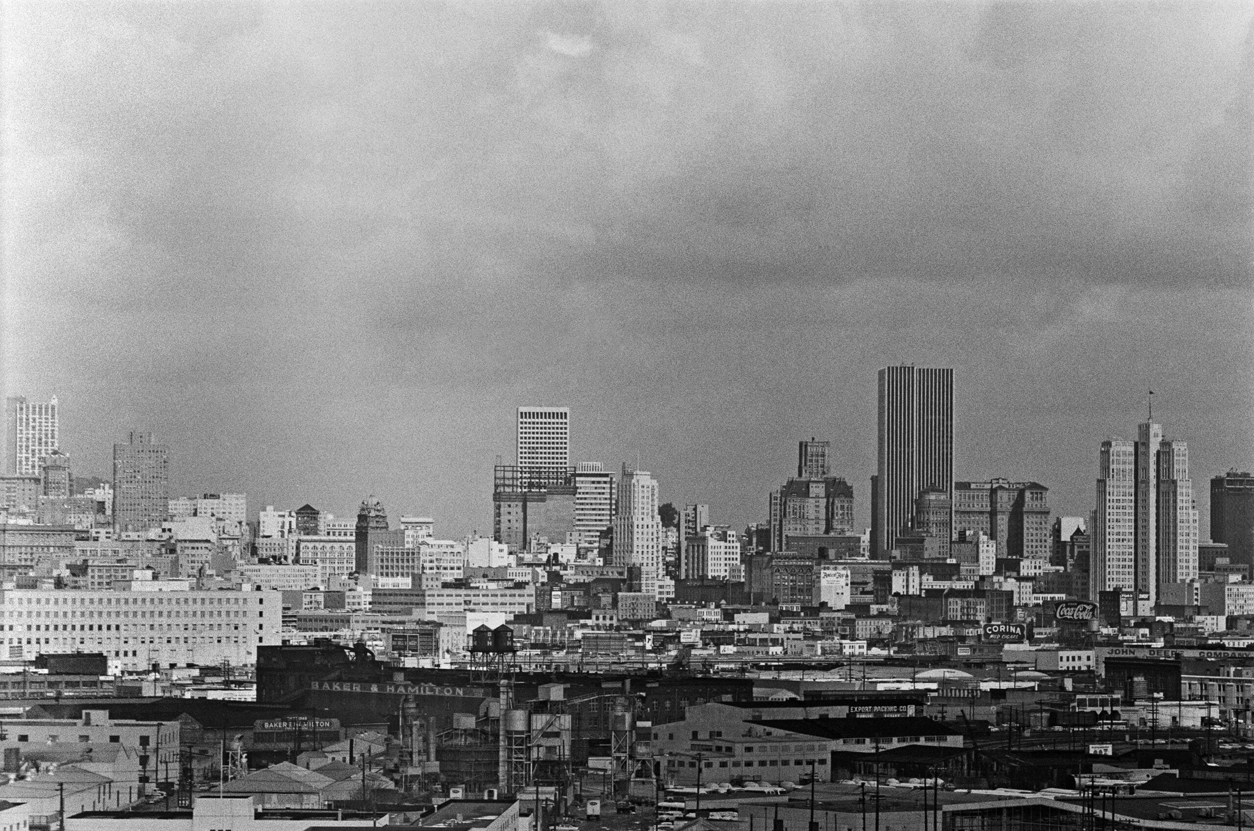 Black and white photo of a city skyline with tall buildings under a cloudy sky. Industrial and commercial structures fill the foreground.