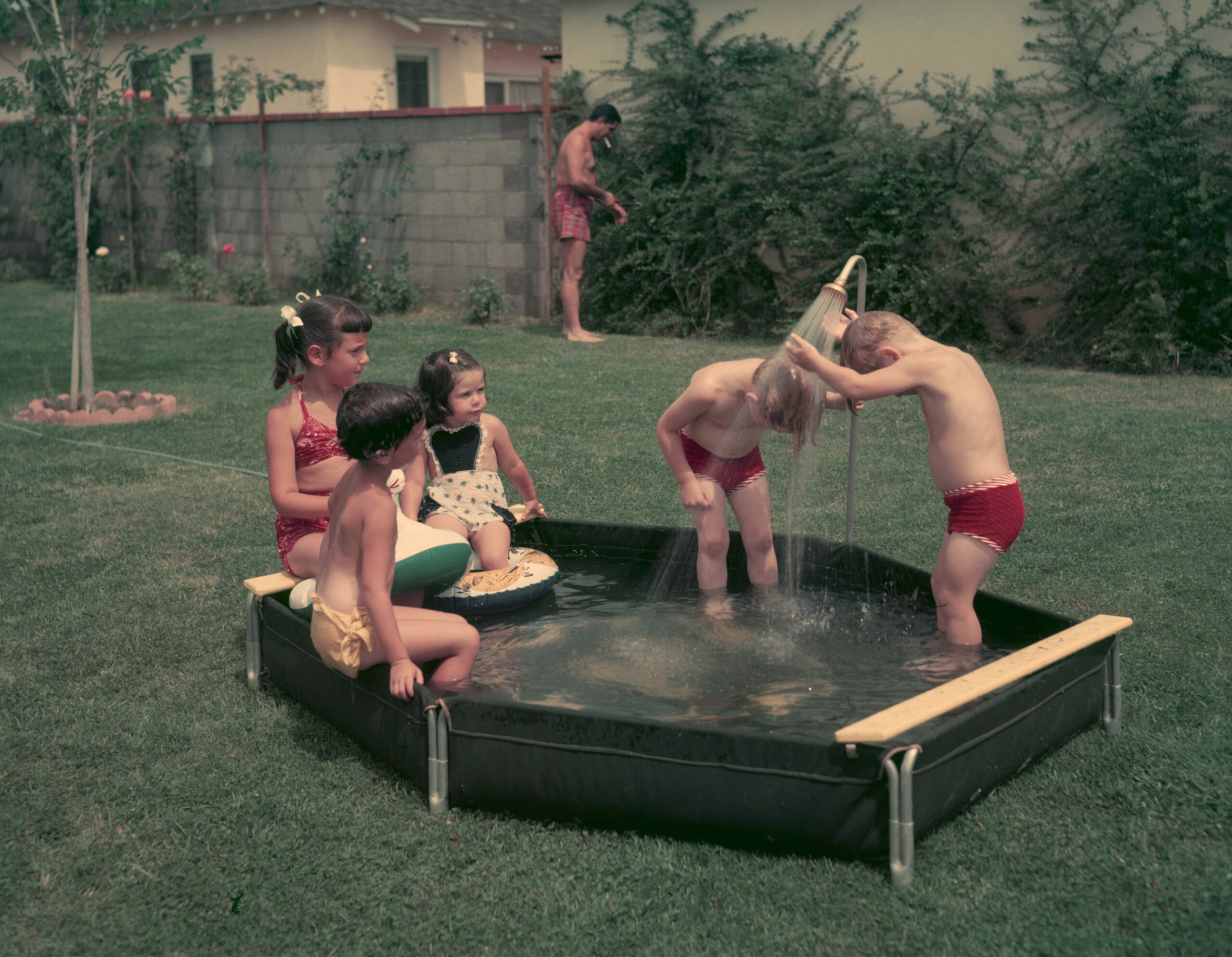 Five young children play in a small backyard pool on a grassy lawn, with one adult standing in the background. Some kids sit at the edge while others stand under a stream of water.