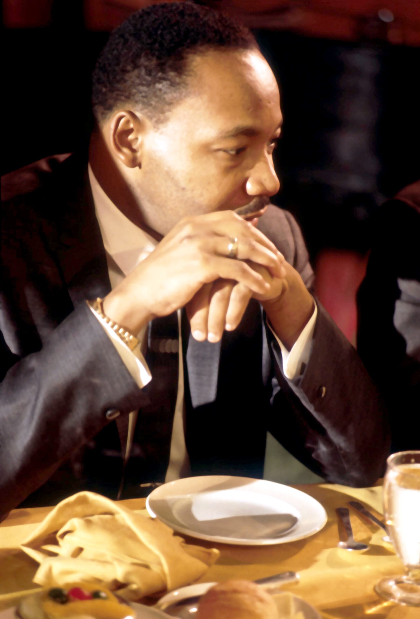 A man in a suit sits at a table with folded hands, looking thoughtful. There is a plate, napkin, and glass in front of him, with warm lighting highlighting his face.