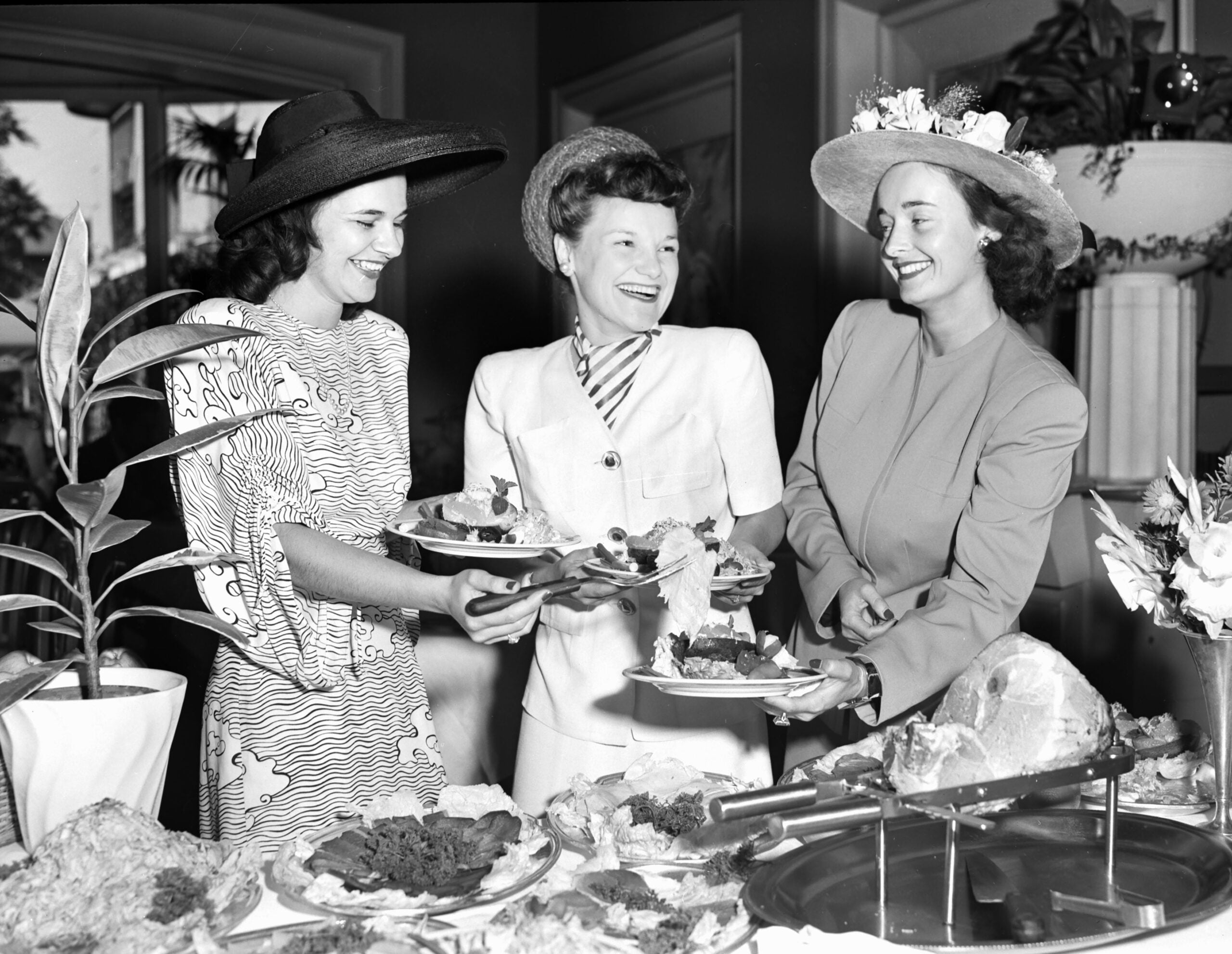 Three women in elegant hats and dresses smile while serving themselves food at a buffet table, surrounded by plates of various dishes and floral arrangements.