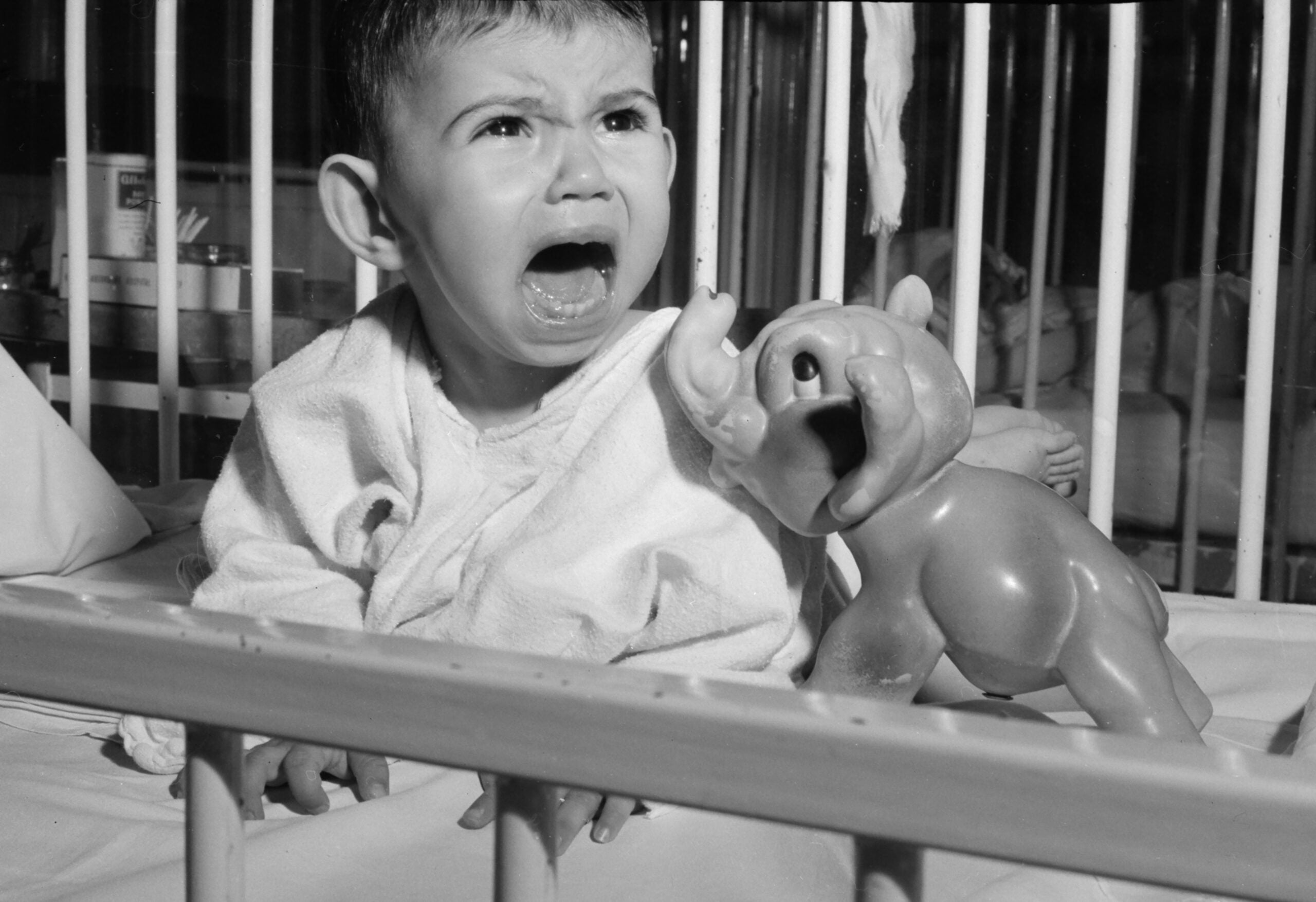 A young child in a crib cries with mouth open, wearing a light gown. A plastic elephant toy sits in front of them. The crib bars are visible in the foreground.