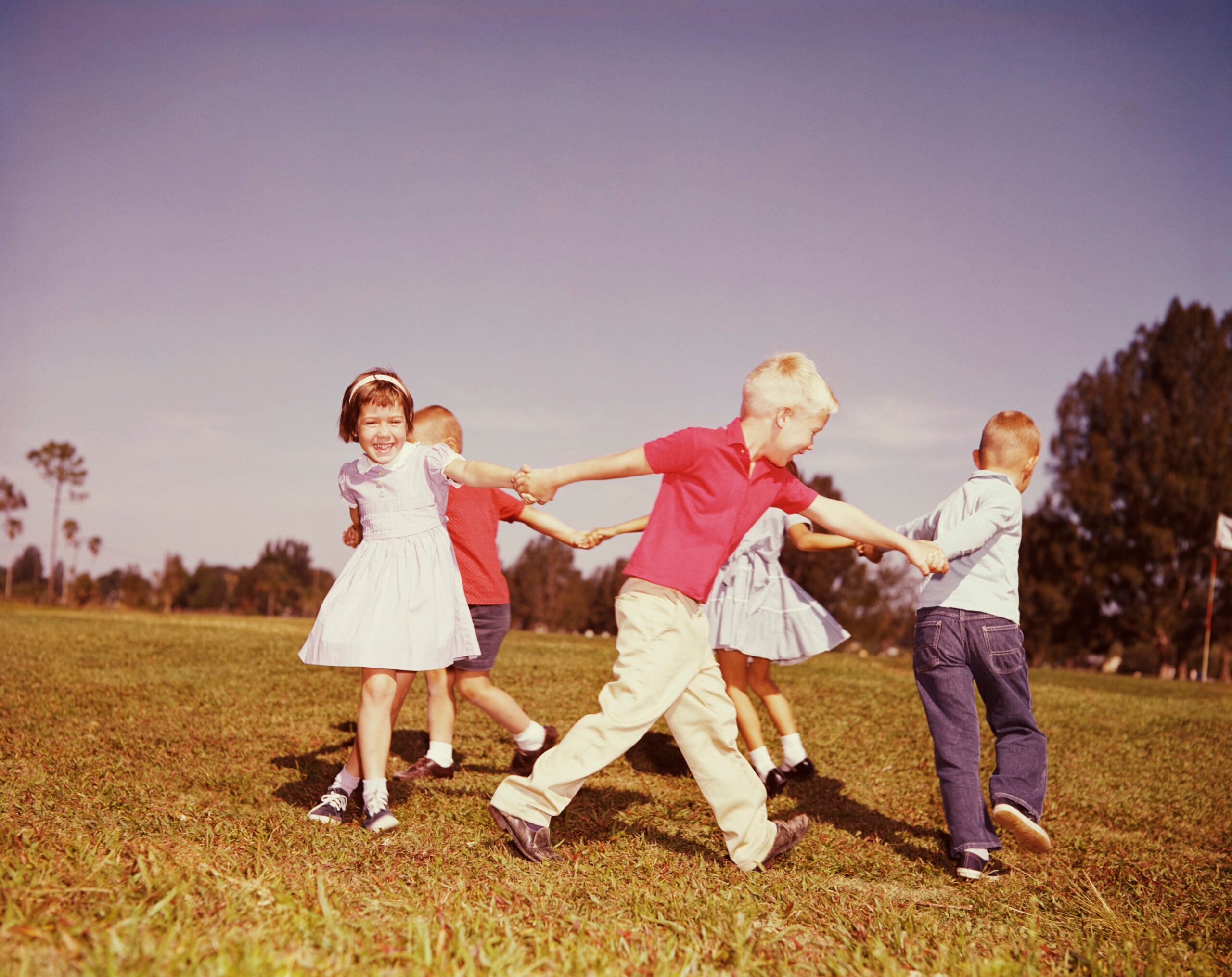 Five young children hold hands and play in a circle on green grass under a clear sky, smiling and laughing, with trees in the background.