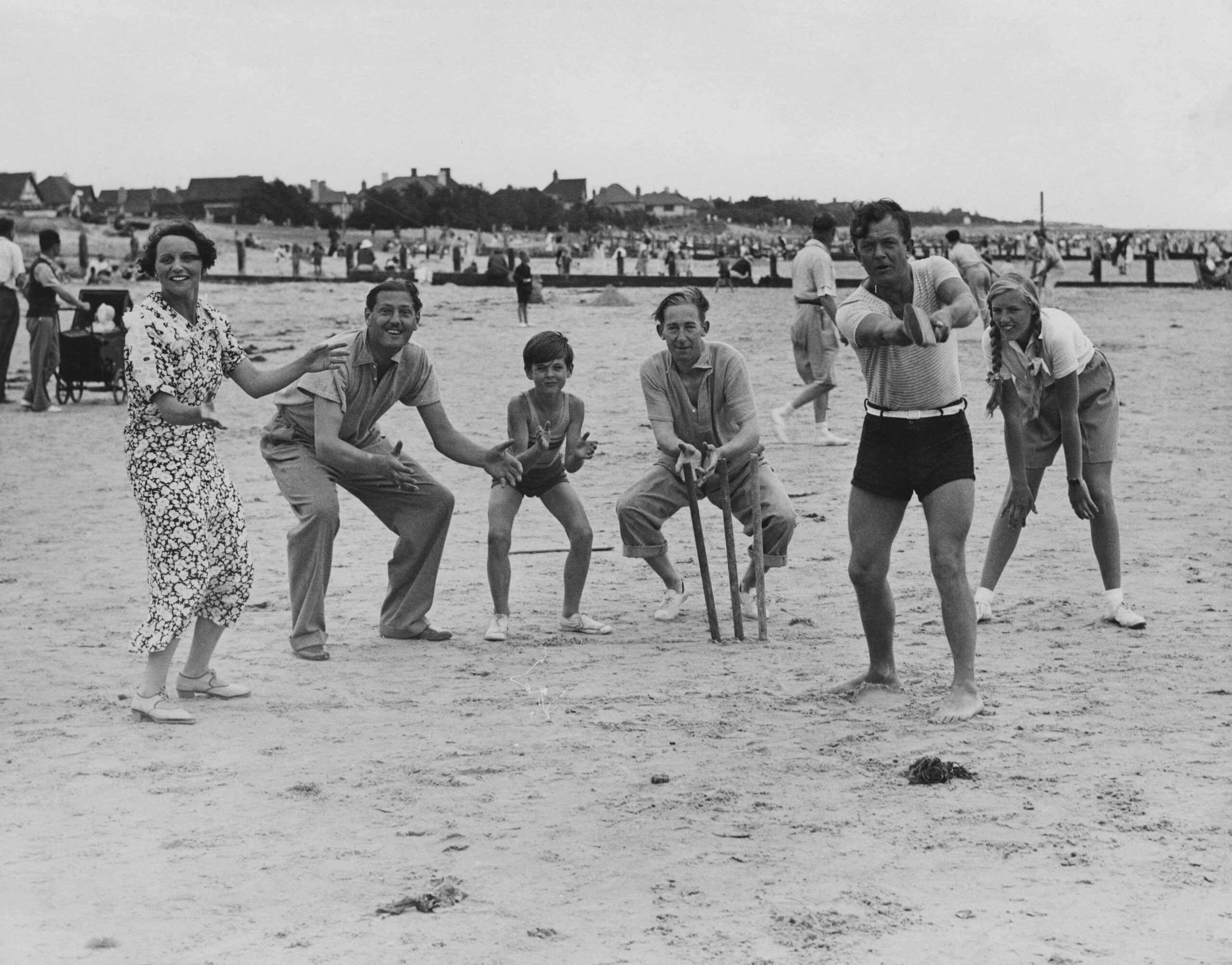 Six people play cricket on a sandy beach, smiling and posing for the camera. Other people and buildings can be seen in the background. The scene appears lively and relaxed.
