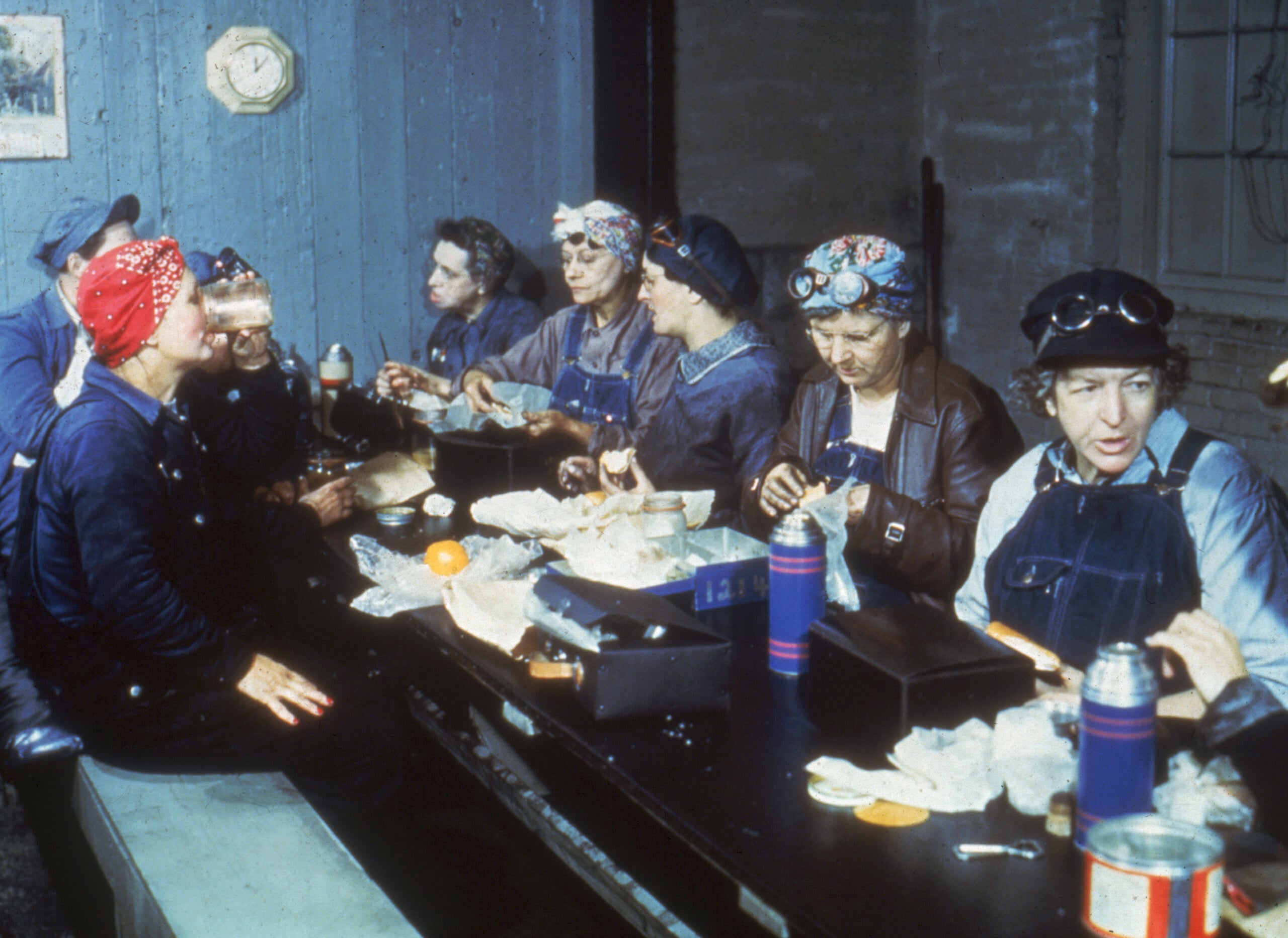Eight women in work clothes and headscarves sit at a table eating lunch, with thermoses and packed food, in a break room with blue walls and a clock on the wall.