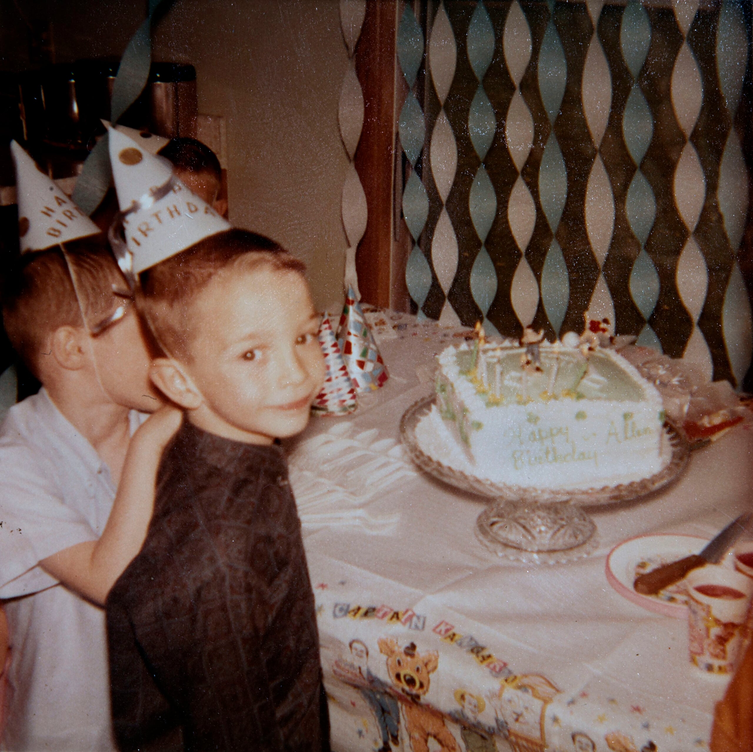 Two boys wearing birthday hats stand by a table with a cake that says “Happy Birthday Alan.” The table is decorated with a cartoon tablecloth, party hats, and plates. Blue and white streamers hang in the background.