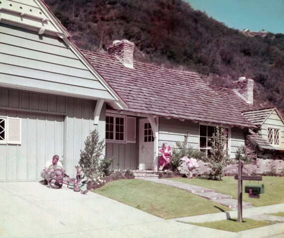 A family is outside a single-story house. Two adults and a child sit by the driveway; another woman sits on the porch. The house has wood siding, a shingle roof, and is set against a hillside.