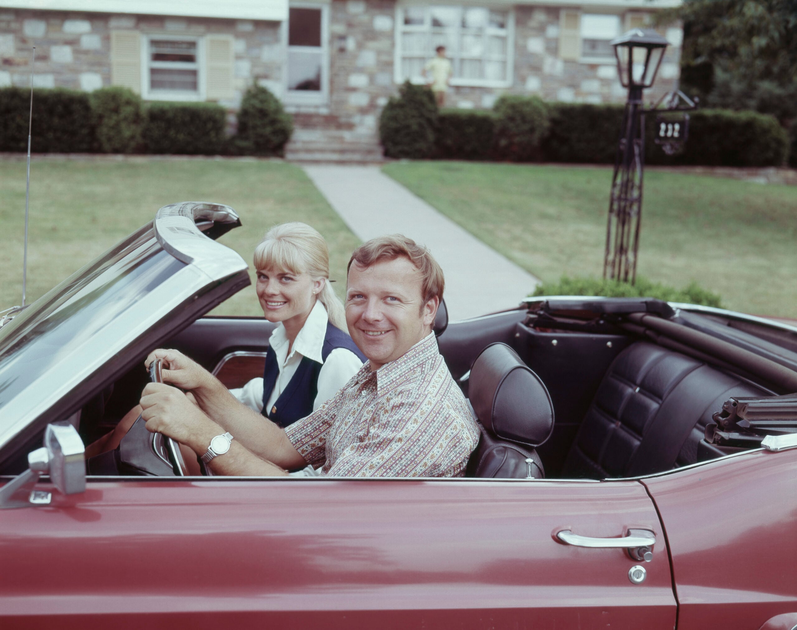 A man and woman smiling while sitting in a red convertible car parked in front of a house with a lawn and stone facade.