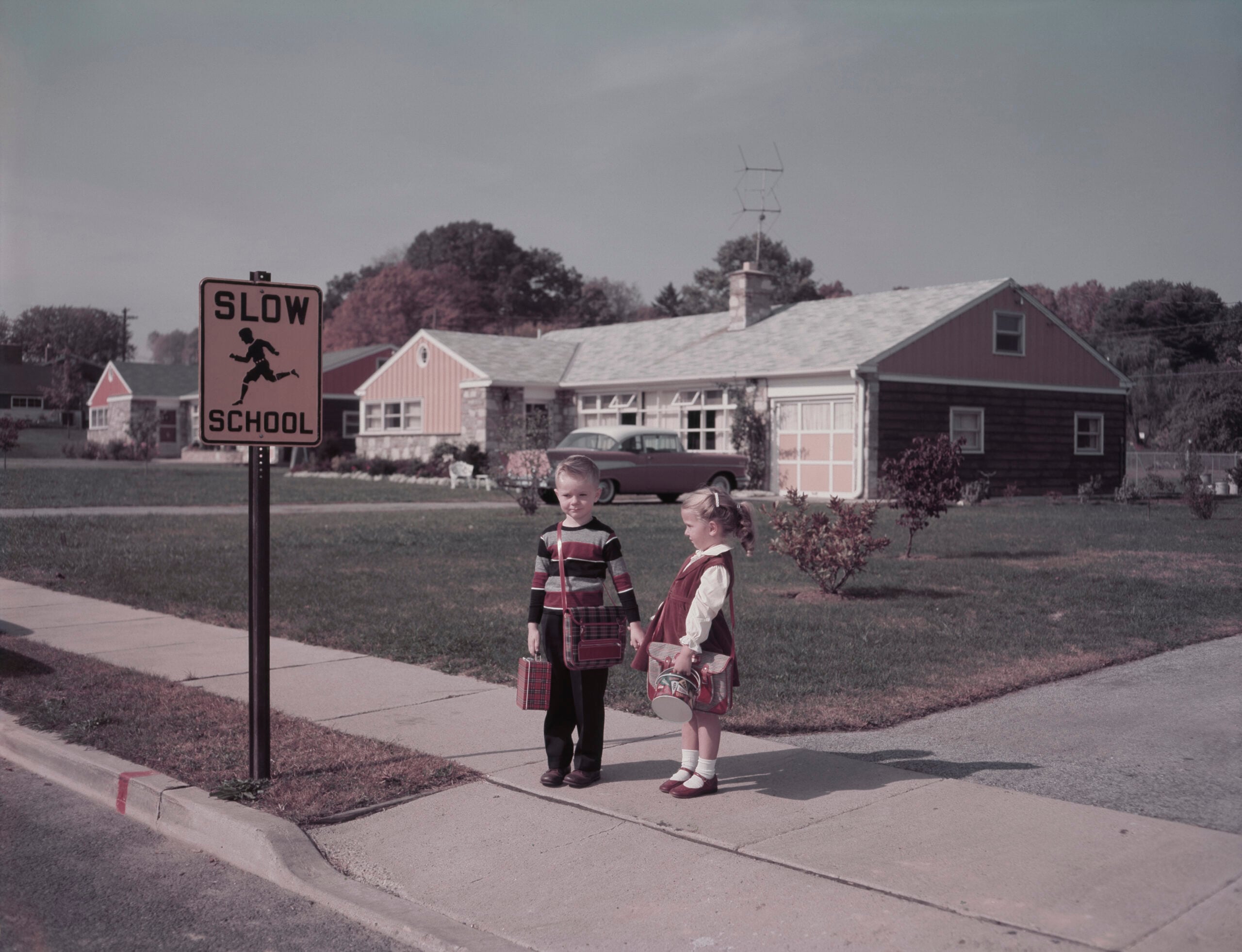 Two young children stand on a sidewalk near a “SLOW SCHOOL” sign, holding lunchboxes, with suburban houses and a vintage car in the background.