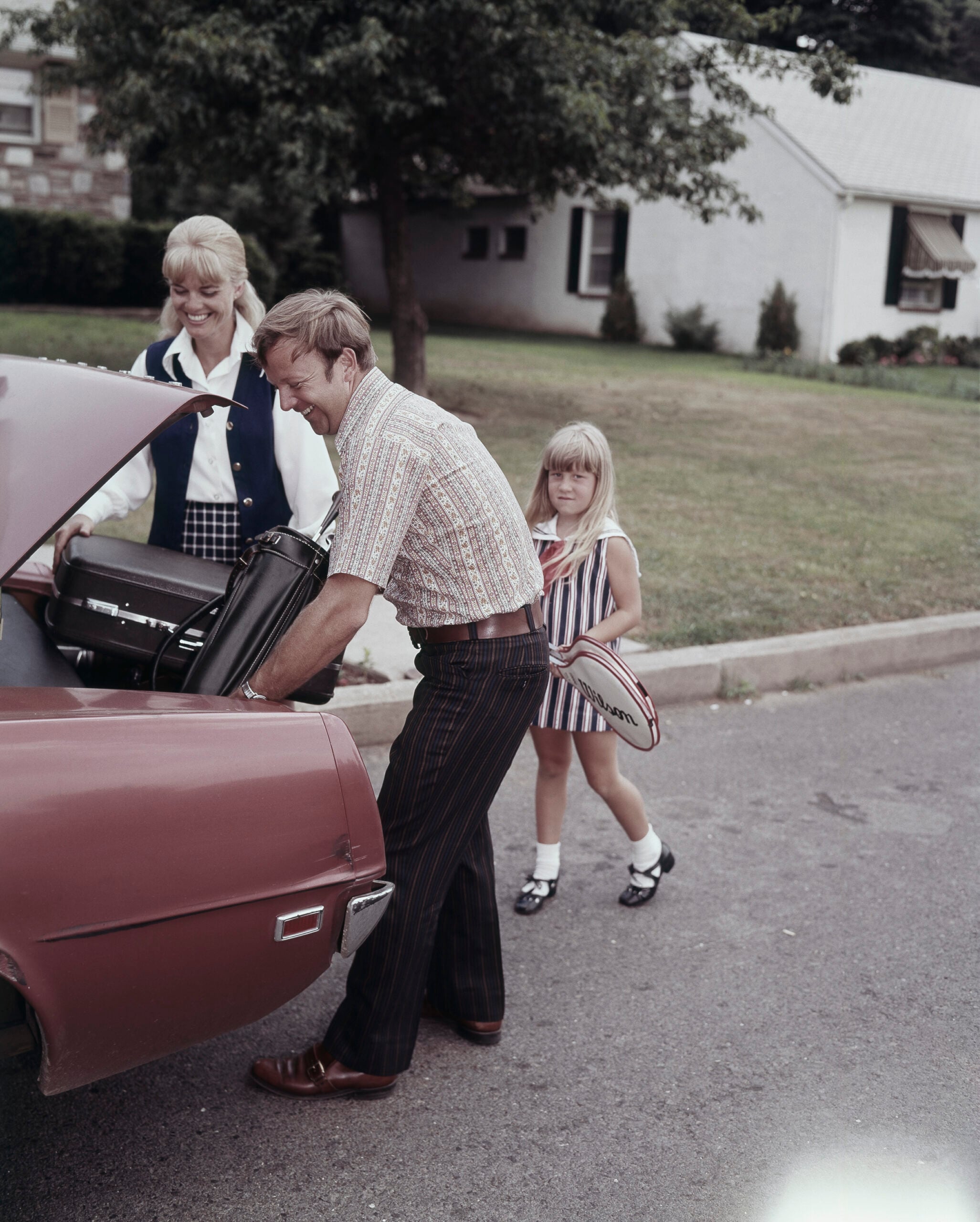 A man loads a suitcase into a red car’s trunk as a woman smiles nearby and a young girl in a striped dress holding a tennis racket stands beside them on a suburban street.