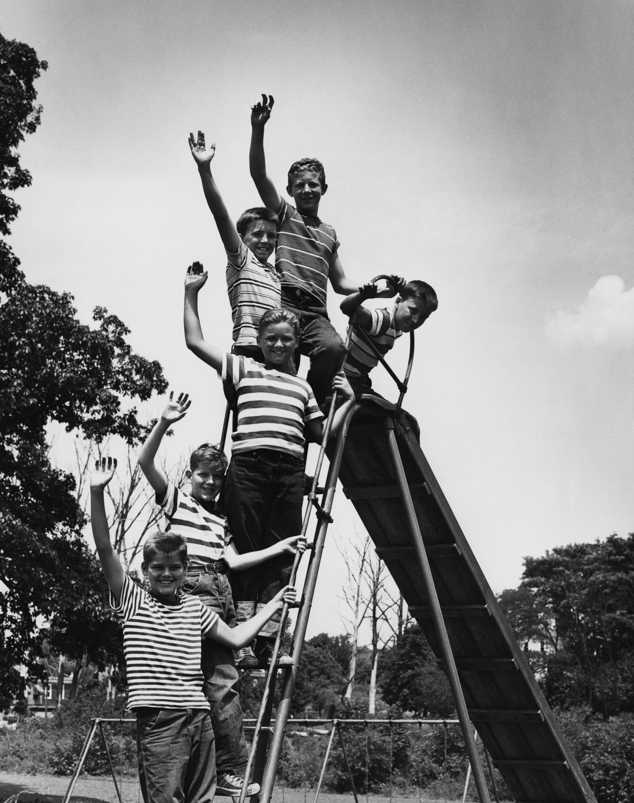 Six children wearing striped shirts stand and wave from the steps and top of a playground slide on a sunny day, with trees and swings visible in the background.