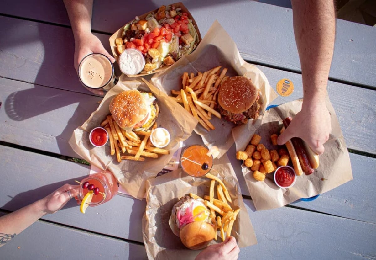 Overhead view of three burgers with fries, a tray of tater tots, two pink drinks, a stout beer, and a loaded salad on a wooden table with three hands reaching for food and drinks.