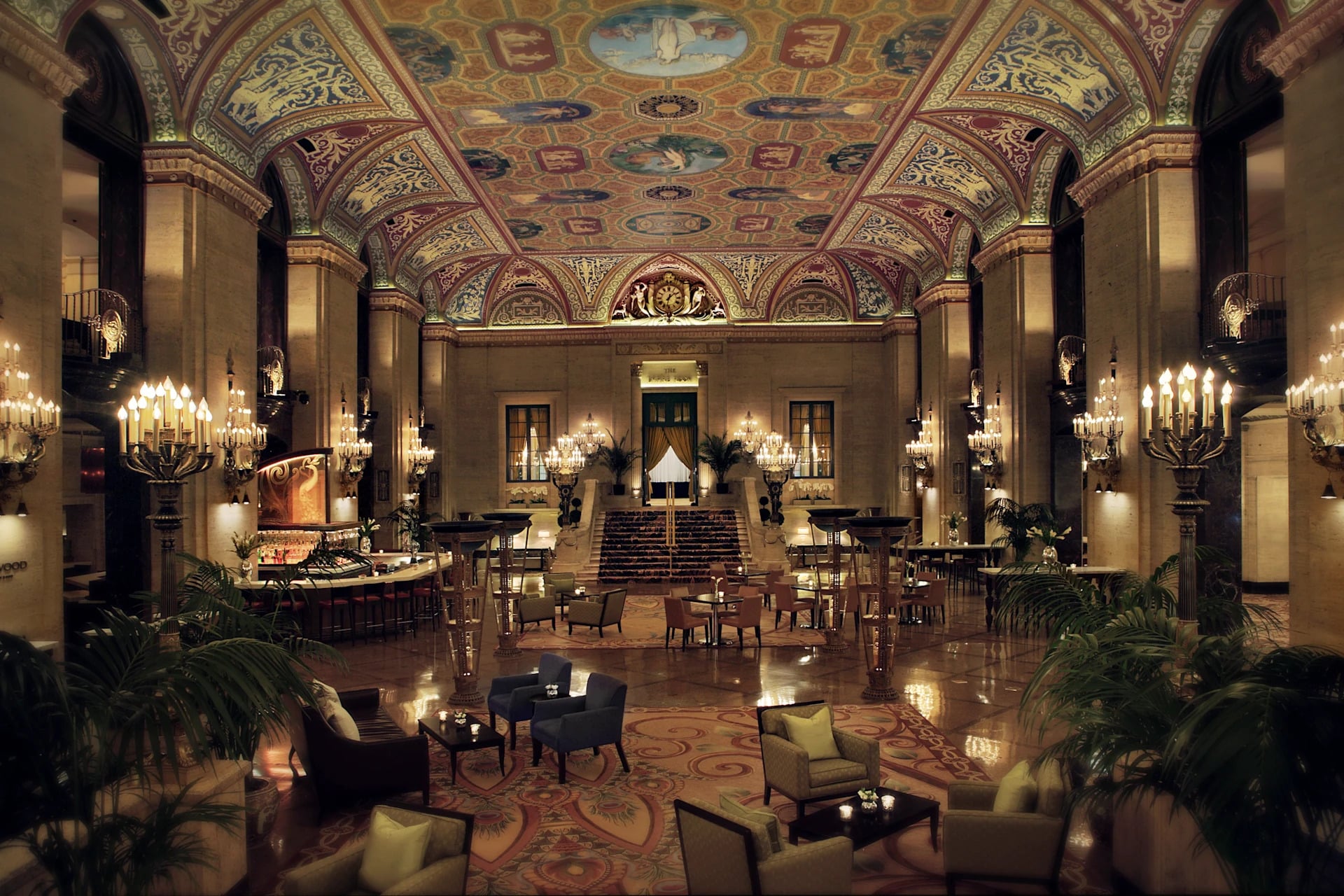 Grand hotel lobby with ornate, colorful vaulted ceiling, elegant chandeliers, plush seating areas, patterned rug, and marble floors. Reflecting the timeless charm of Historic Hotels, stairs lead up to gold-trimmed doors surrounded by plants and warm lighting.