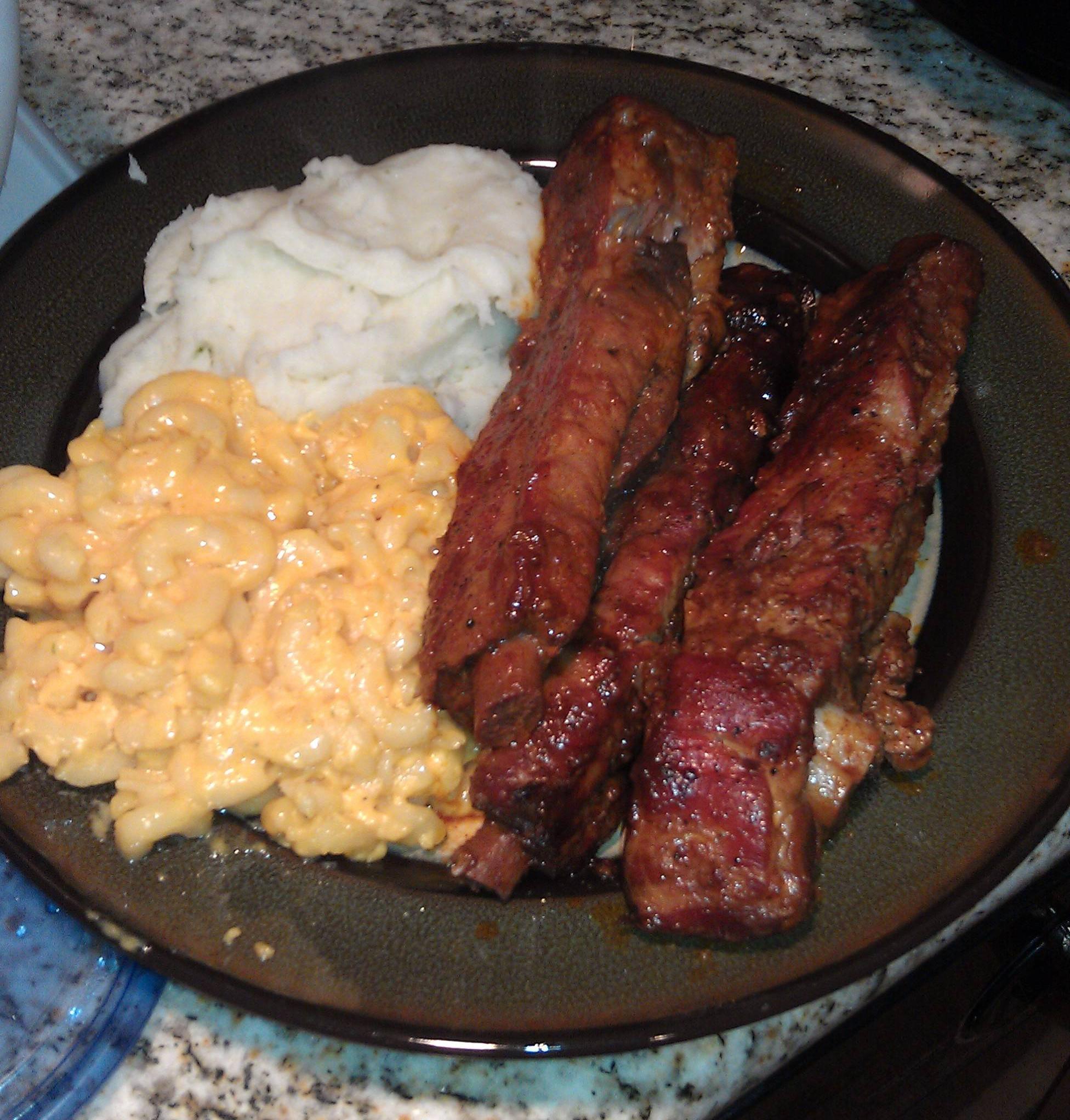 A plate with three ribs, a serving of macaroni and cheese, and a portion of mashed potatoes, set on a granite countertop.