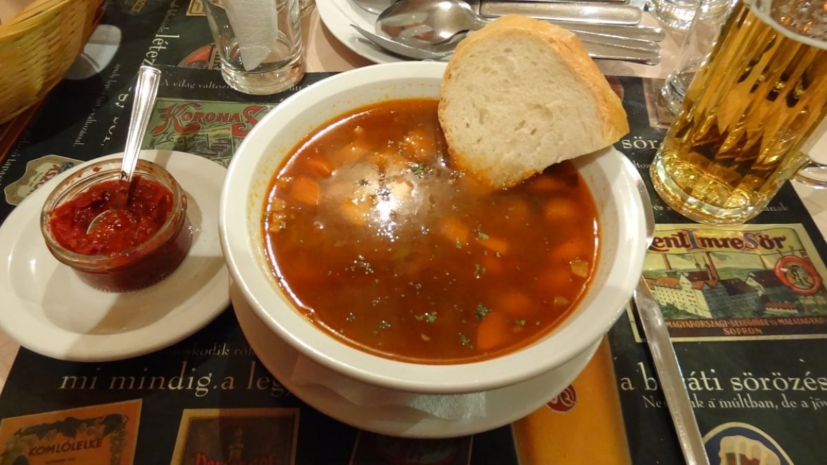 A bowl of red soup with vegetables, topped with chopped herbs and a slice of bread, sits on a table next to a glass of beer and a small dish of red sauce with a spoon—truly an example of the amazing food people ate while on vacation.