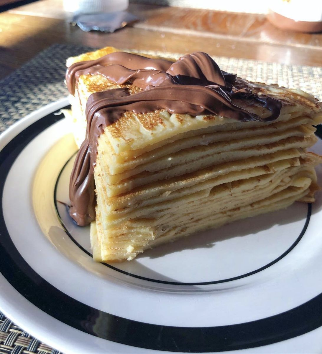 A slice of crepe cake with multiple thin layers, topped with a generous swirl of chocolate spread, sits on a white plate with black trim. Sunlight highlights the dessert.