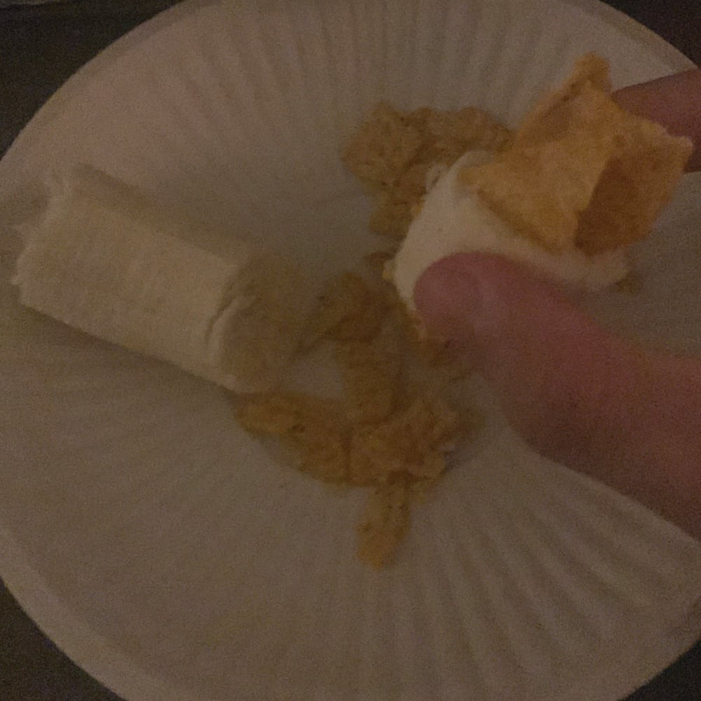 A hand holds a banana slice with cereal flakes pressed into it over a white paper plate containing more banana and cereal pieces.