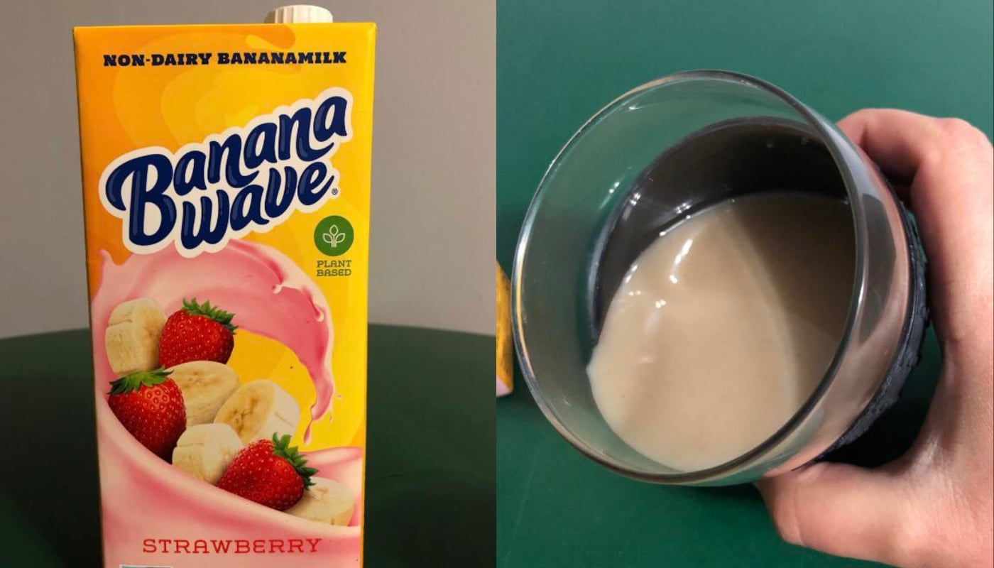 A carton of Banana Wave strawberry non-dairy banana milk sits next to a hand holding a glass filled with the creamy pink beverage, on a green surface.