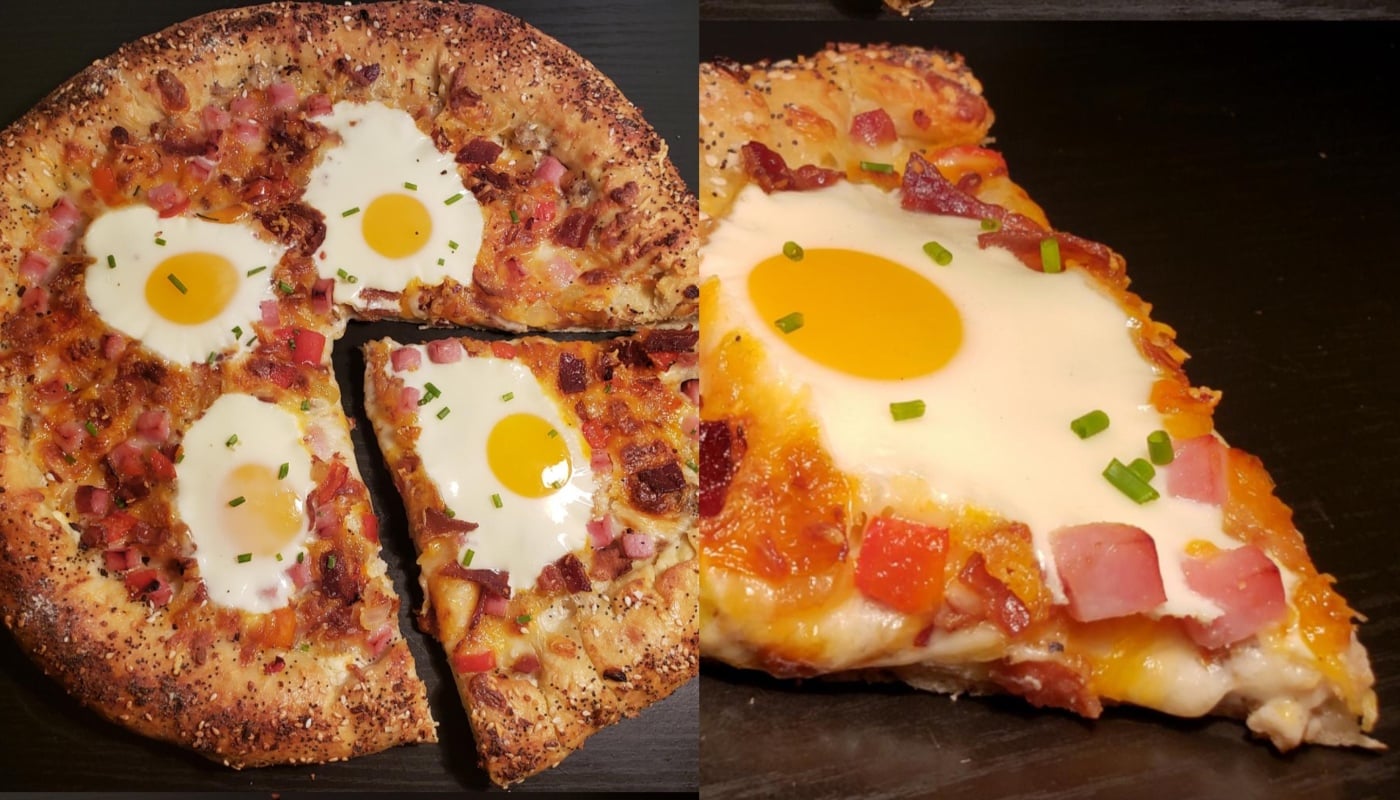 A pizza with a seeded crust, topped with cheese, diced ham, bacon, and whole baked eggs. One slice is separated, showing melted cheese and a runny egg yolk, garnished with chopped chives.