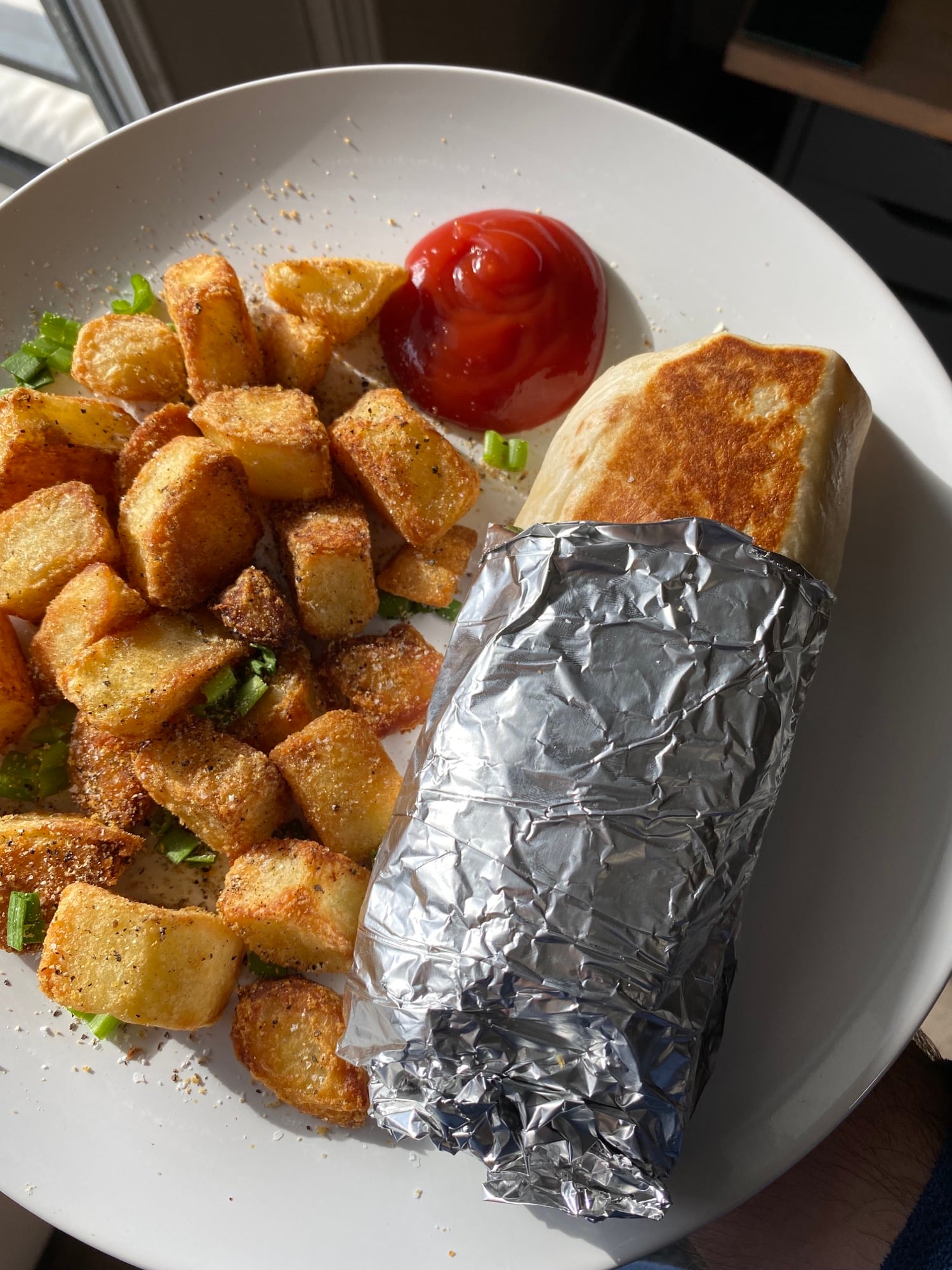 A plate with seasoned diced potatoes, a dollop of ketchup, and a foil-wrapped burrito on a sunny table.