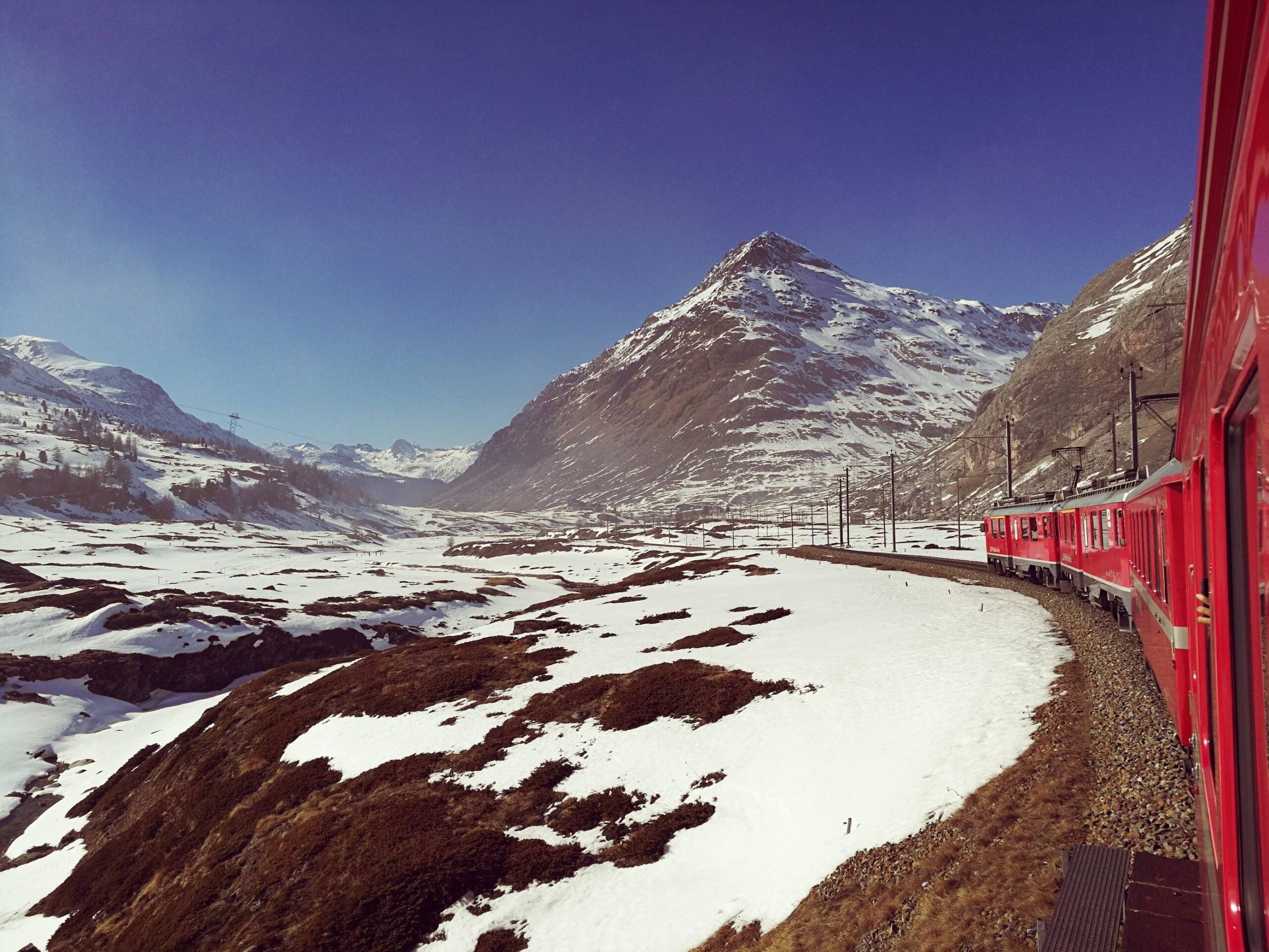 A red train travels through a snowy mountain landscape under a clear blue sky, curving along tracks with snow-capped peaks and brown patches of earth visible.
