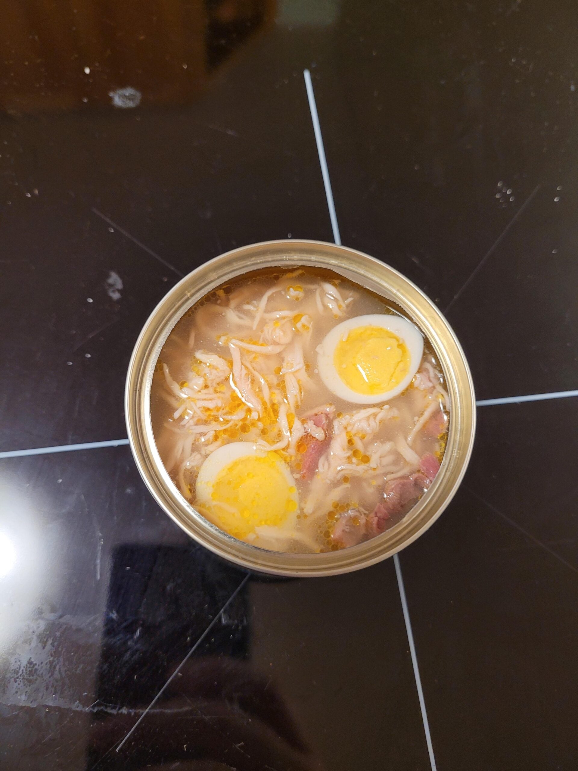 An opened can of soup with noodles, meat, and halved boiled eggs on a dark reflective surface.
