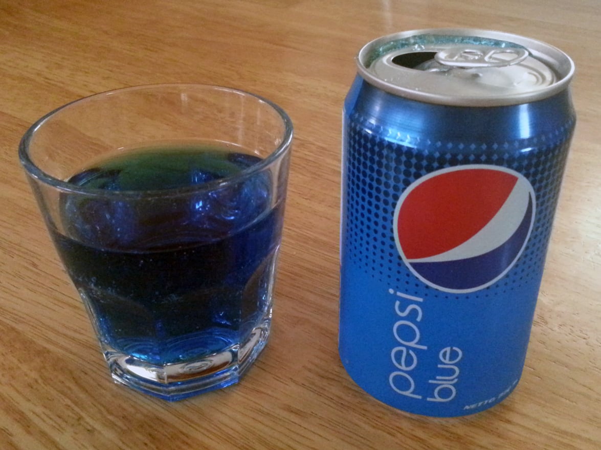 A glass filled with blue soda pops sits next to an opened can of Pepsi Blue on a wooden table.