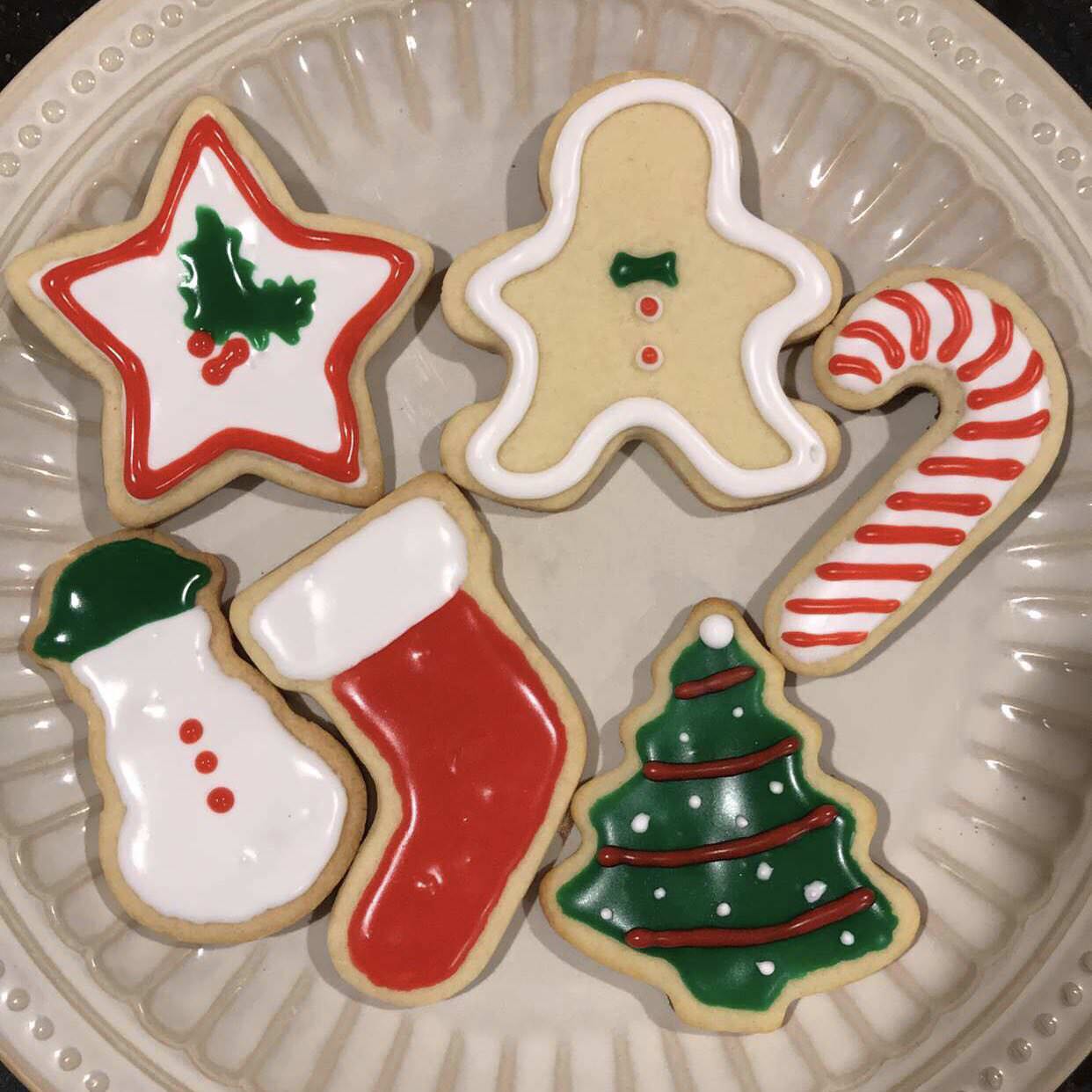 Six decorated homemade Christmas treats sit on a plate: a red and white star, gingerbread person, candy cane, snowman, red stocking, and green Christmas tree.