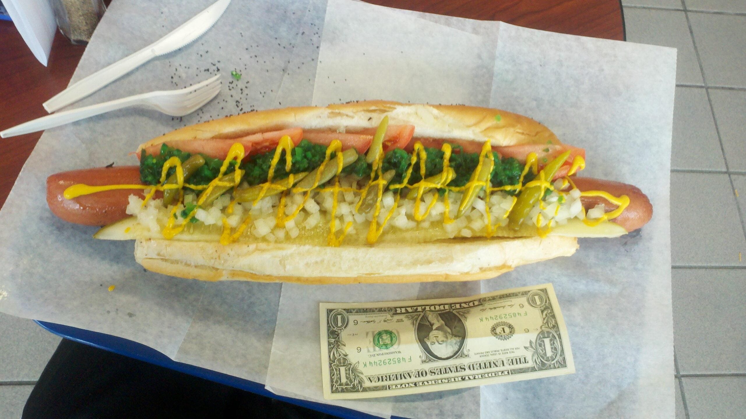 A large hot dog in a bun with onions, relish, tomato, and mustard sits on parchment paper next to a U.S. dollar bill and two plastic forks on a table.