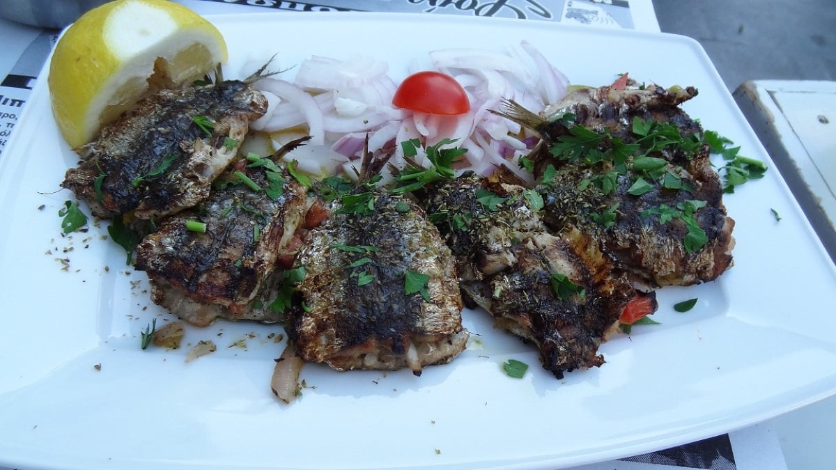 Grilled sardines garnished with chopped herbs, served with sliced red onions, a lemon wedge, and a cherry tomato on a white rectangular plate—the kind of amazing food people ate while on vacation, bringing back delicious memories of travel.