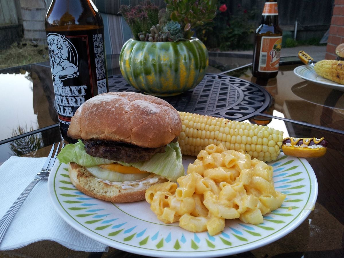 A plate with a cheeseburger, macaroni and cheese, and corn on the cob on an outdoor table, with beer bottles, a napkin, and a green pumpkin-shaped planter in the background.