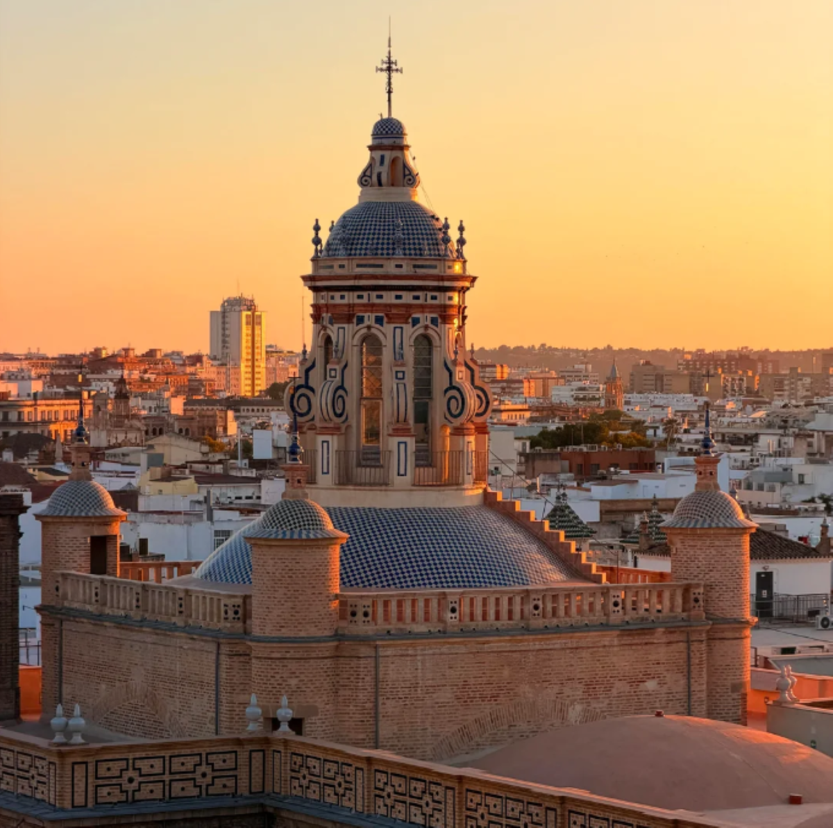 A historic domed building with ornate details stands amid a cityscape at sunset, with warm sunlight illuminating rooftops and distant modern buildings under an orange sky.