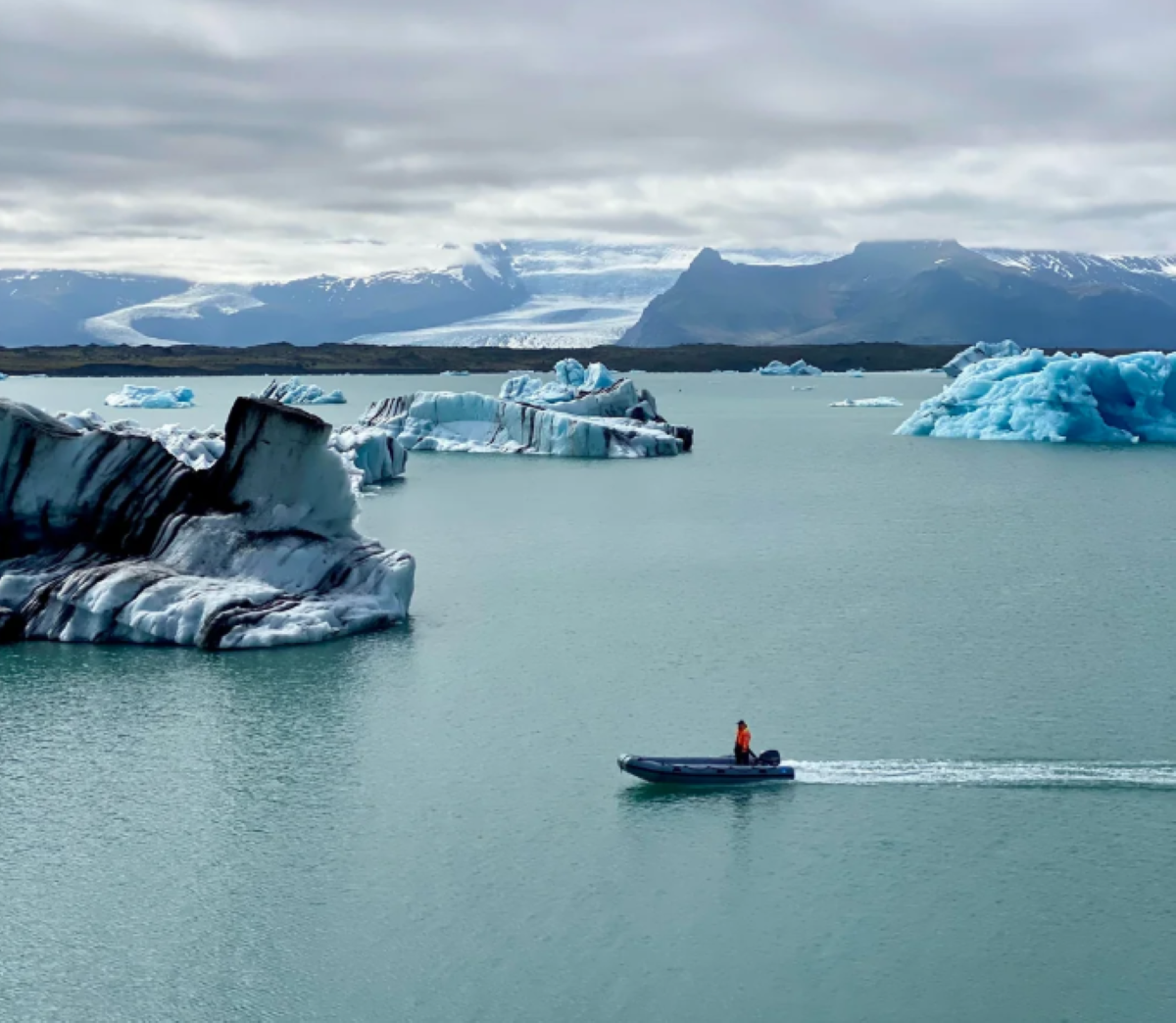 A person in a small boat travels across icy water surrounded by large blue icebergs, with snowy mountains and a glacier in the background under a cloudy sky.