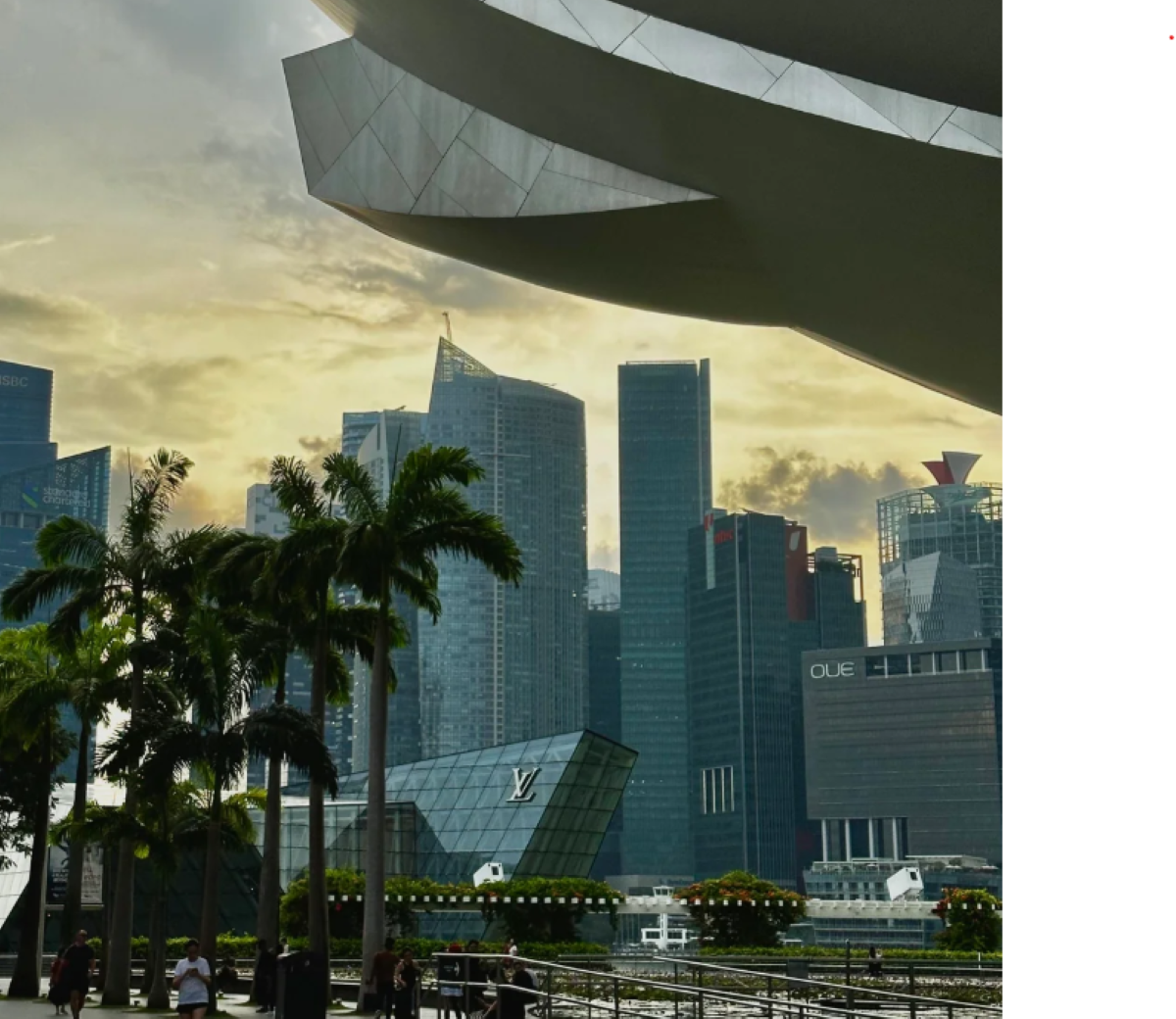 Tall modern skyscrapers, palm trees, and unique architecture line a waterfront promenade at sunset, with dramatic clouds and a few people walking in the foreground.