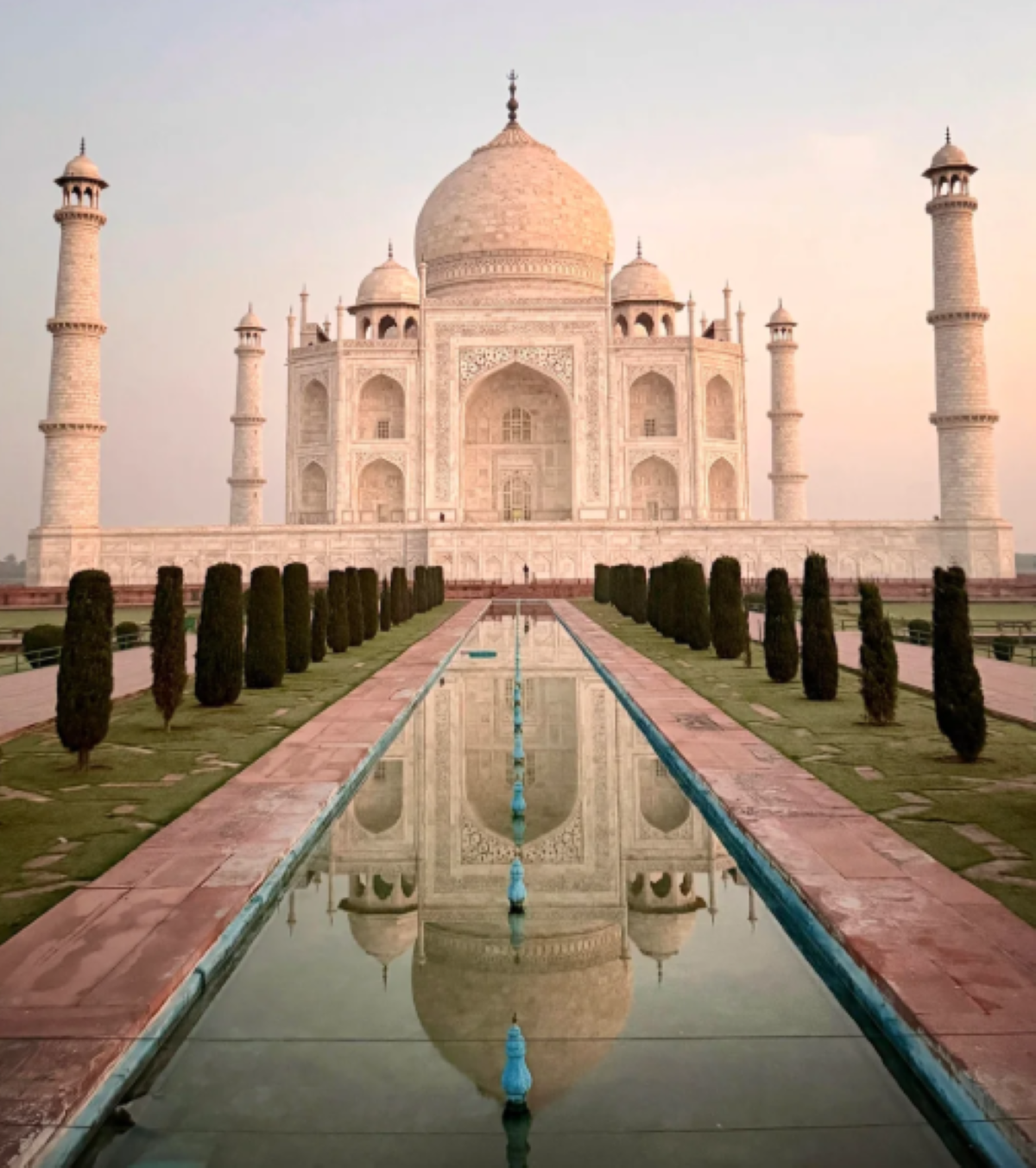 The Taj Mahal at sunrise, its white marble facade reflected in a long rectangular pool, flanked by trimmed bushes and pathways under a clear sky—an iconic sight often featured among travel experiences that went viral.