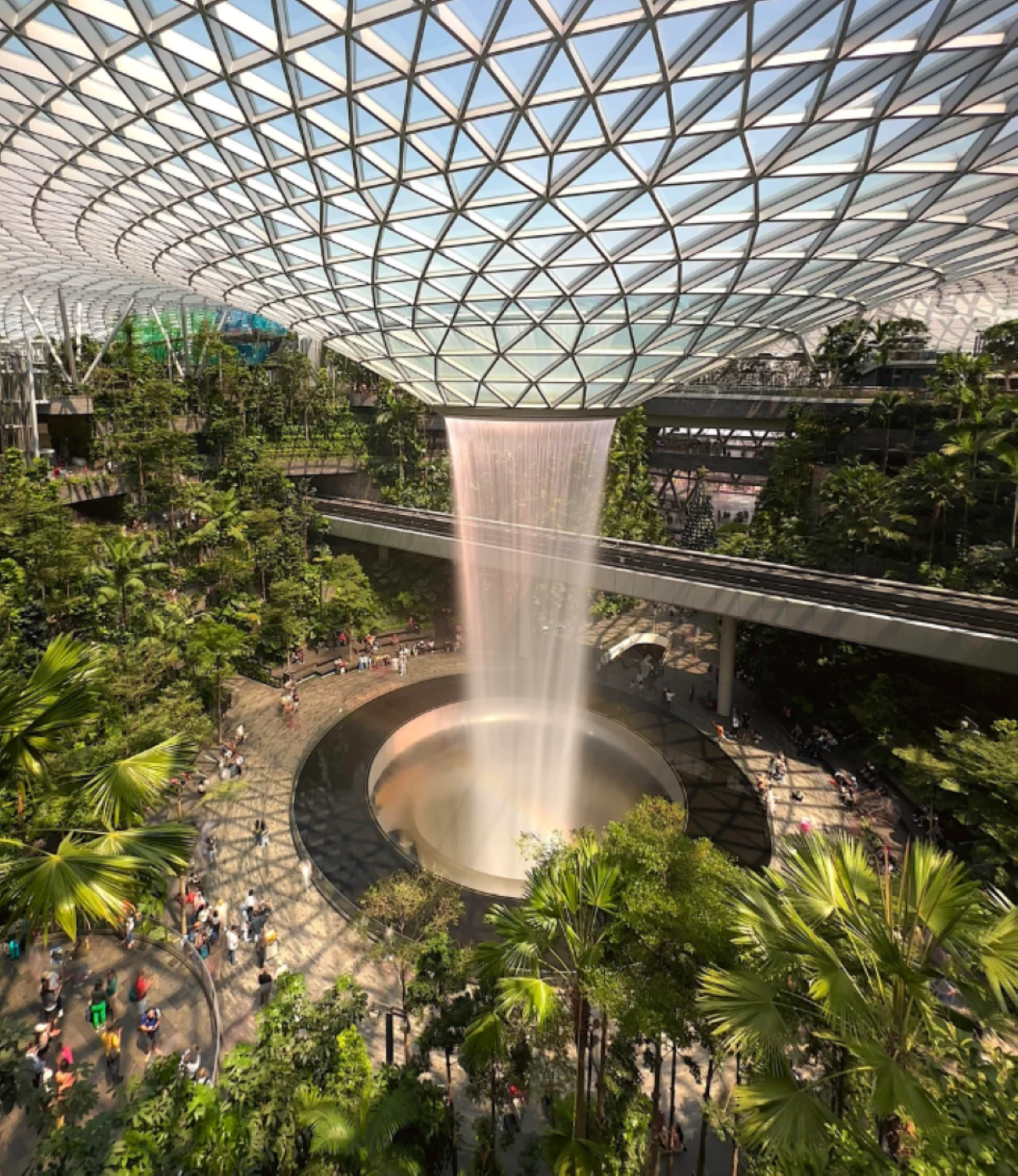 A large indoor waterfall, featured in travel experiences that went viral, cascades into a circular pool surrounded by lush greenery and people, all beneath a striking geometric glass dome ceiling.