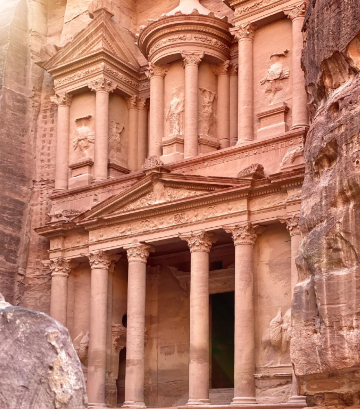 The image shows the ancient rock-cut facade of Al-Khazneh, also known as The Treasury, in Petra, Jordan, featuring ornate columns and detailed carvings in pink sandstone. Sunlight softly illuminates the structure, capturing the timeless wonder that has made Petra one of those travel experiences that went viral around the world.