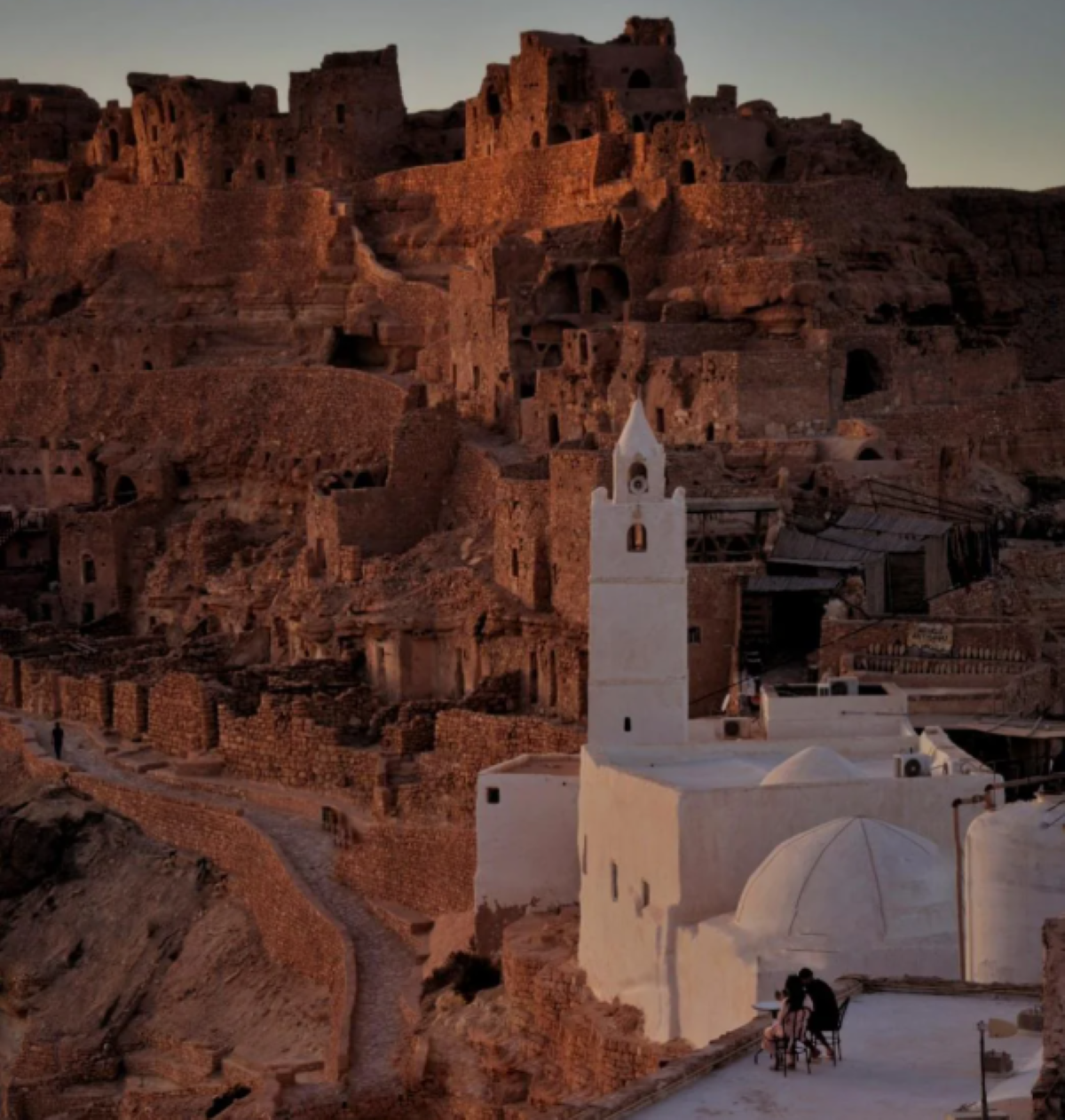 Ancient hilltop ruins glow in sunset light behind a white mosque with a tall minaret. Two people sit together in the foreground, overlooking the dramatic, rugged landscape—a scene reminiscent of travel experiences that went viral for their breathtaking beauty and sense of discovery.