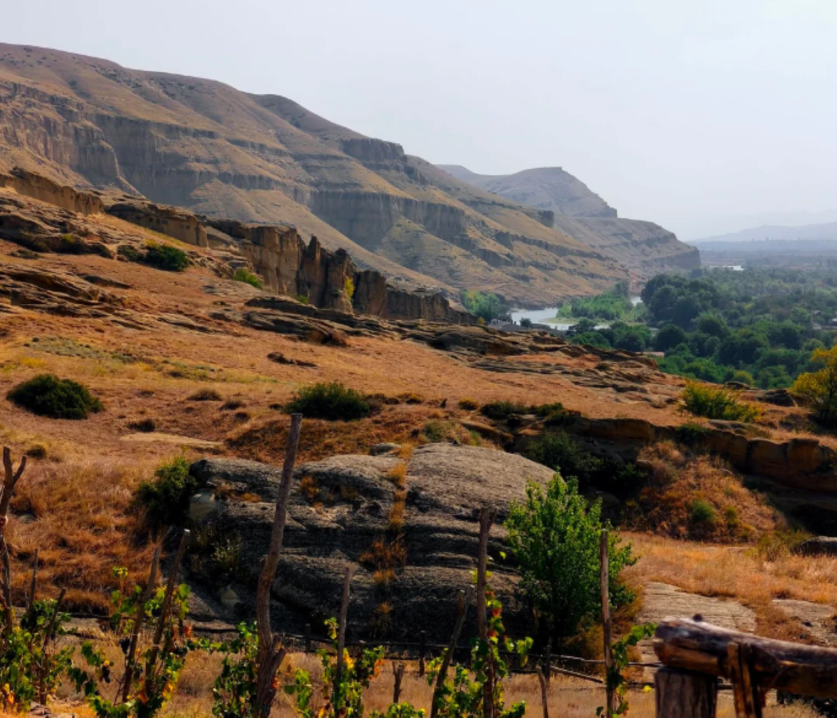Rocky hills with layered cliffs and sparse vegetation overlook a winding river bordered by green trees, under a hazy sky—a landscape reminiscent of travel experiences that went viral for their raw beauty and untouched charm. The foreground features dry grass, shrubs, and a simple wooden fence.