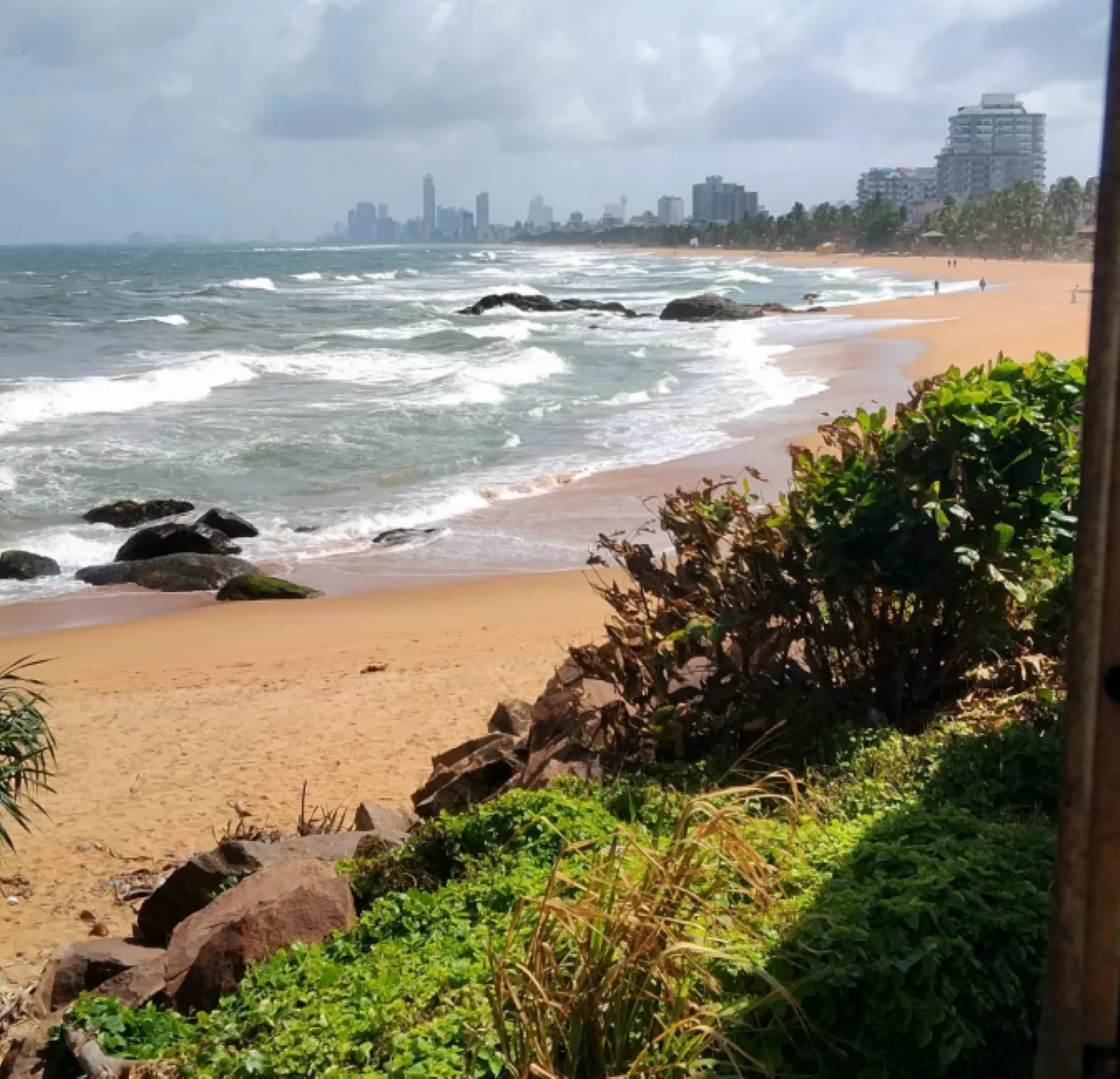 Waves crash on a sandy beach lined with rocks and greenery. Skyscrapers and buildings are visible in the distance along the coastline under a cloudy sky.