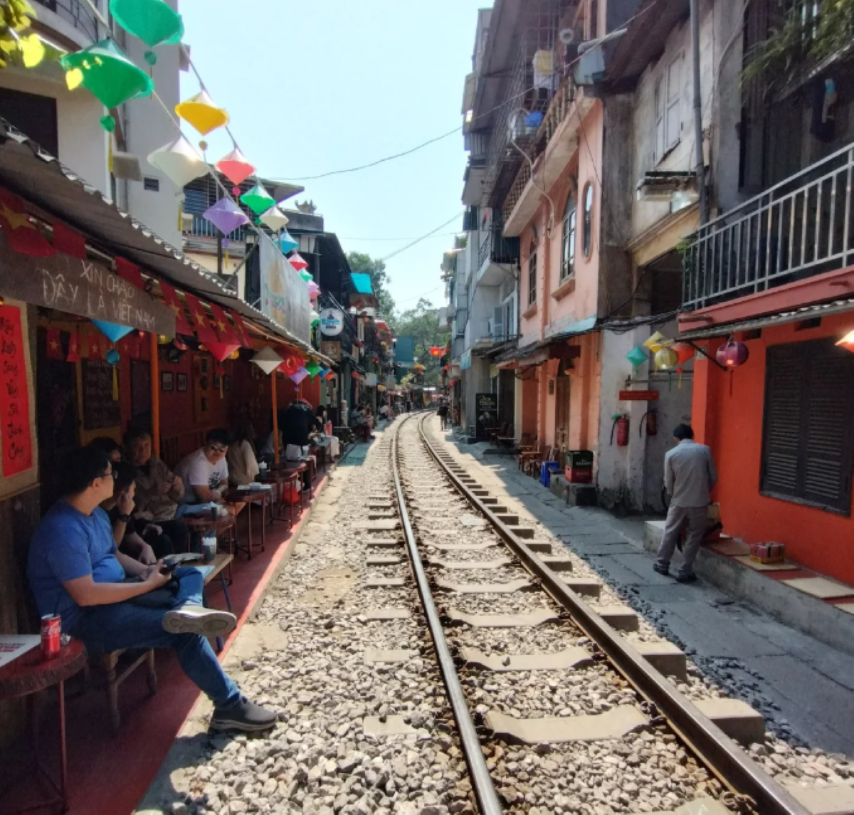 A narrow railway track runs between colorful buildings with hanging lanterns. People sit at small cafes along the tracks, enjoying drinks in the shade. The track curves and disappears into the distance under a sunny sky.