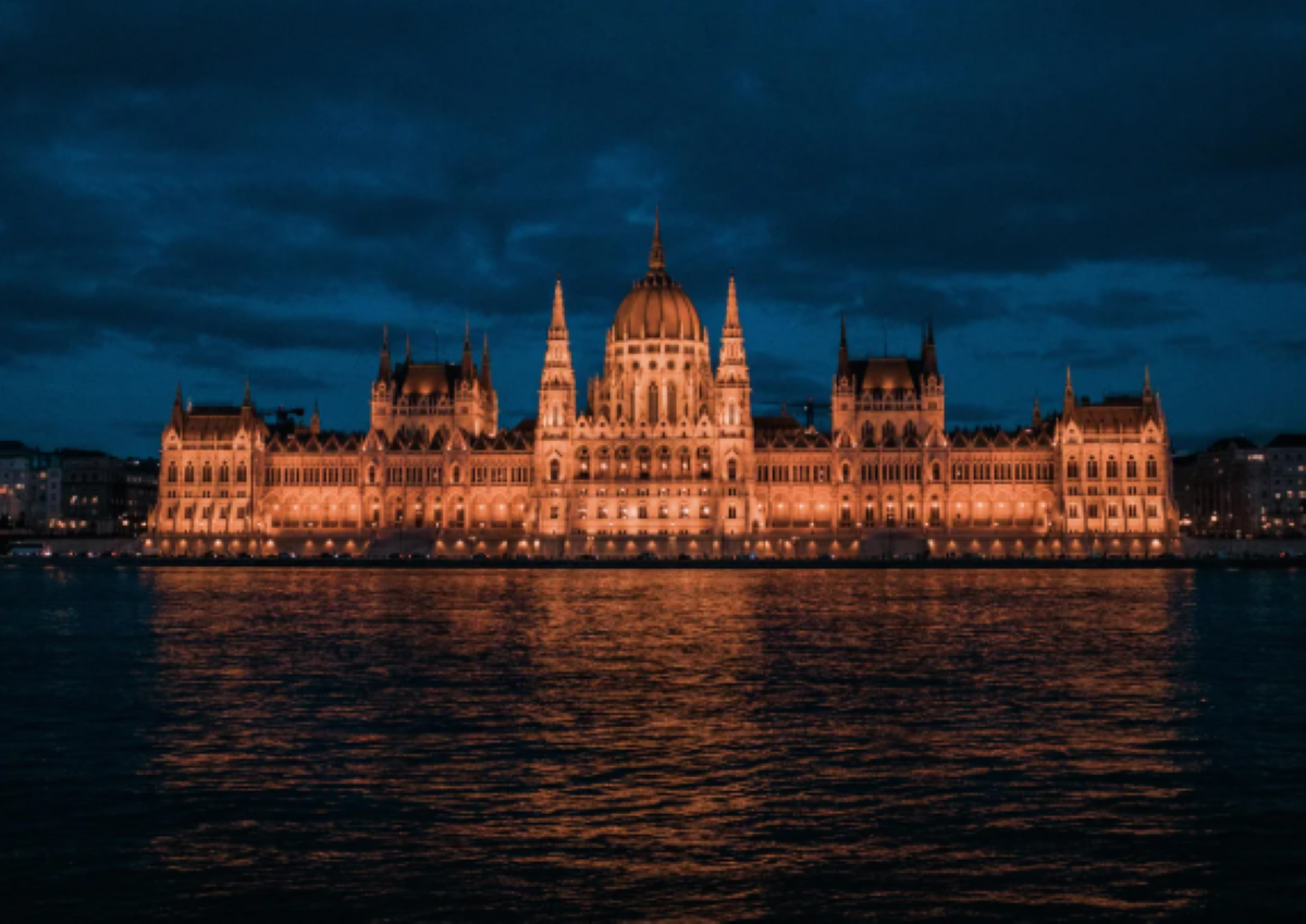 The illuminated Hungarian Parliament Building at night, reflected in the waters of the Danube River under a dark, cloudy sky.